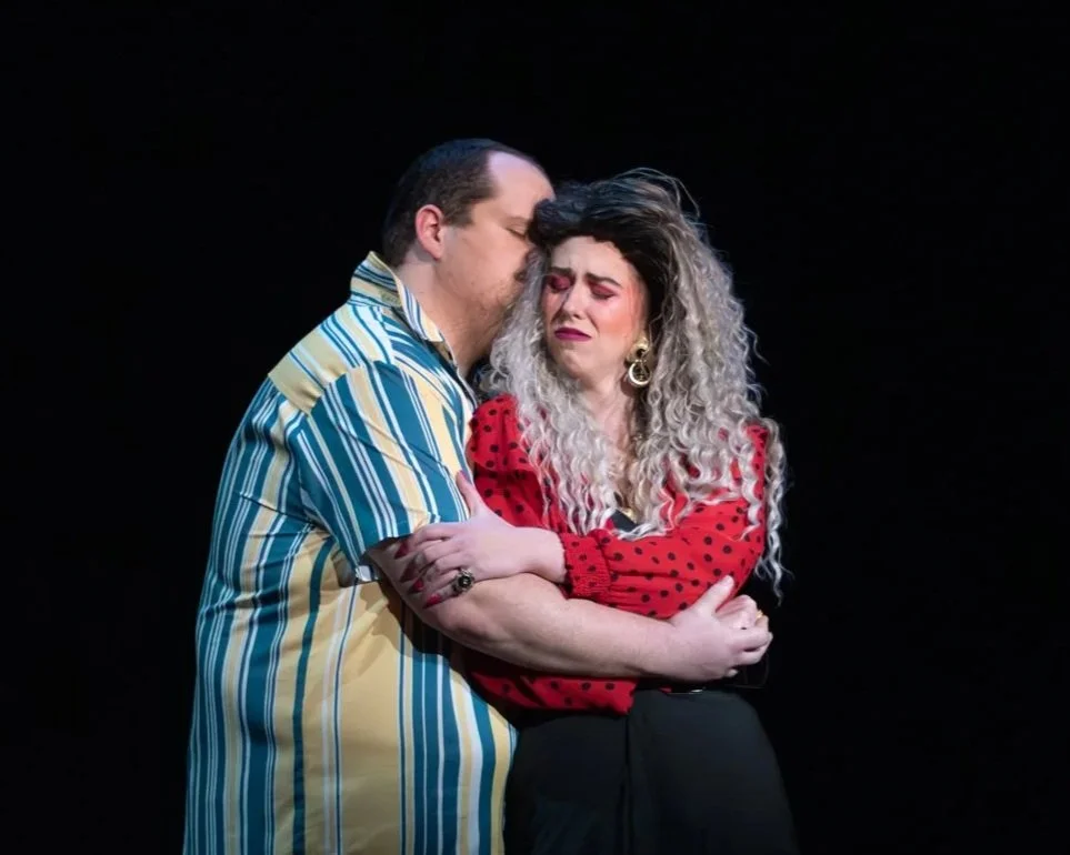 A man and woman on stage, with emotional expressions, sharing an intimate moment. The man has short dark receding hair and the woman has curly hair with blonde and gray streaks, wearing bold makeup and earrings.