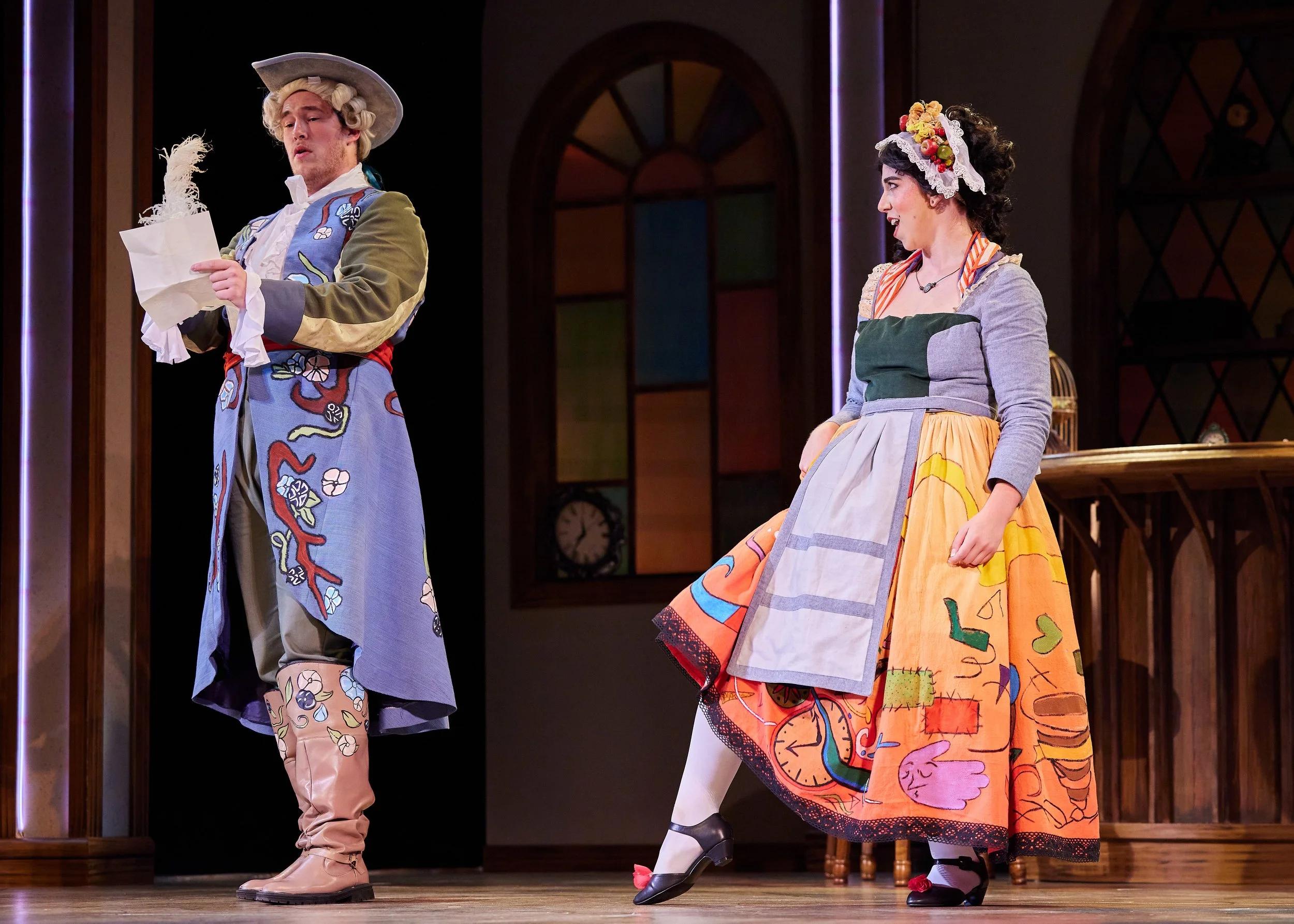 Two actors on stage wearing colorful, historical costumes. The male actor on the left is reading a letter, wearing a large powdered wig, a long blue coat with floral embroidery, beige trousers, and tall beige boots. The female actor on the right is s