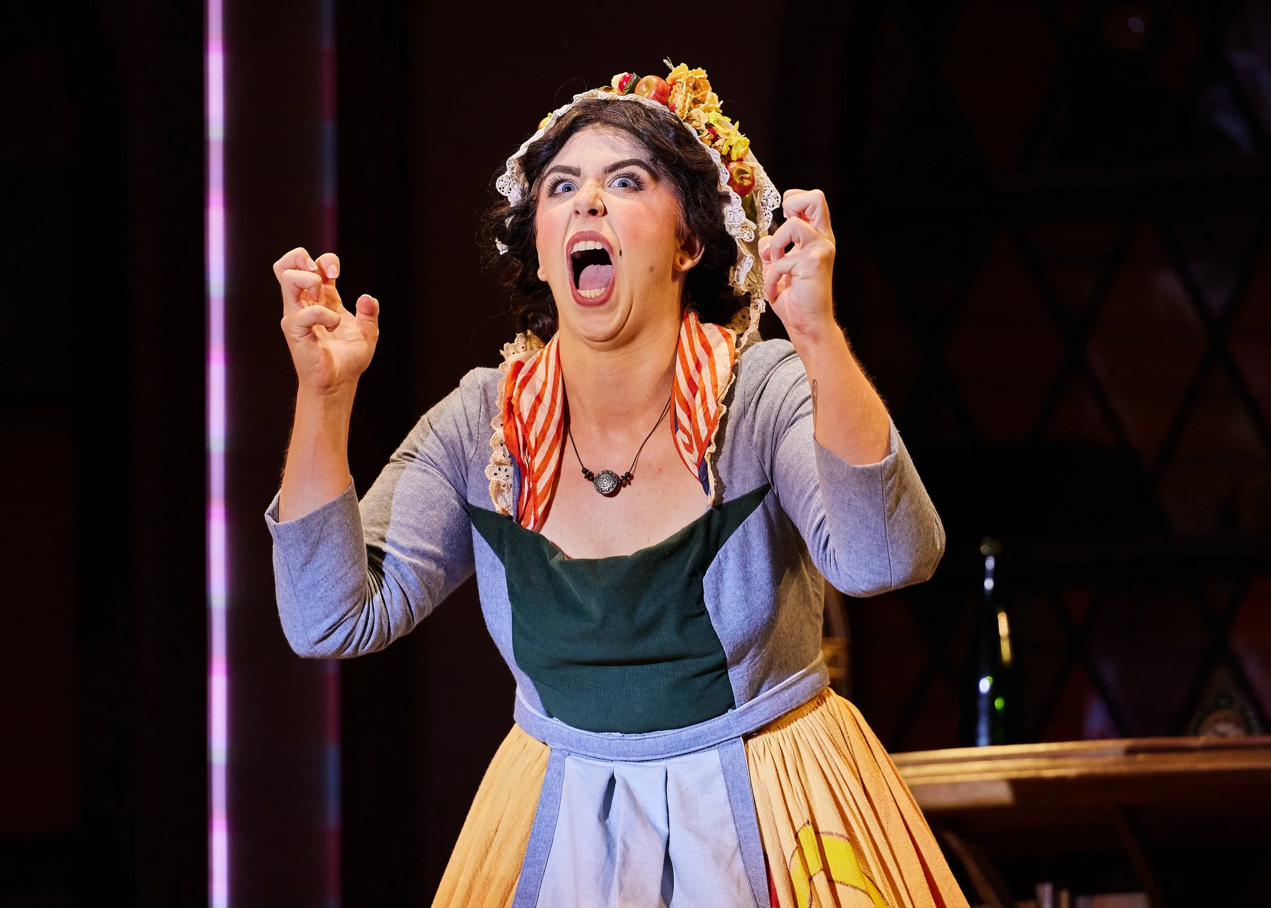 A woman in traditional dress with a apron, wearing a bonnet decorated with flowers, is shouting or singing passionately with raised fists on stage.
