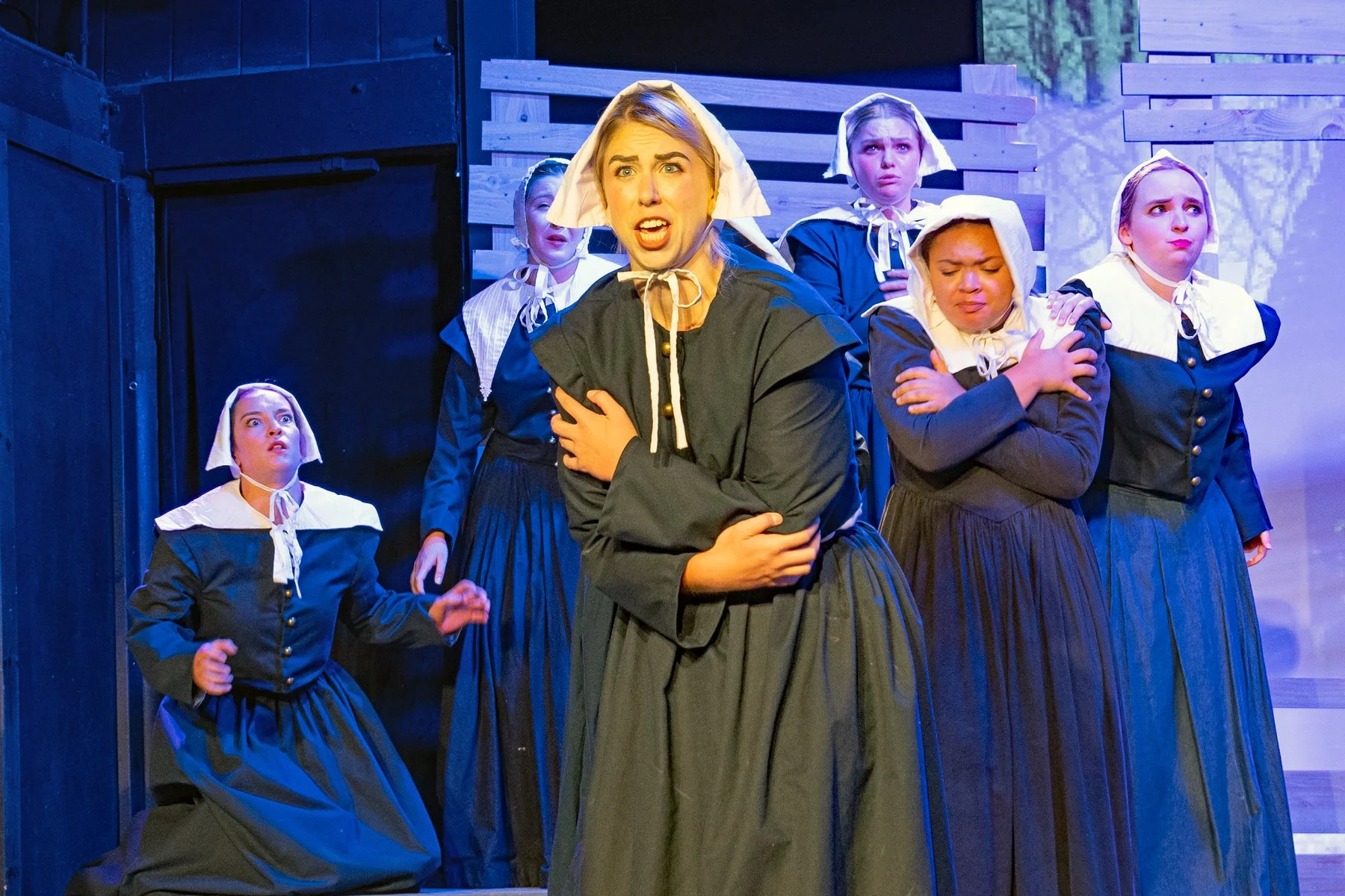Women dressed as Pilgrims performing on stage with expressions of concern or fear, set against a backdrop of wooden scenery.