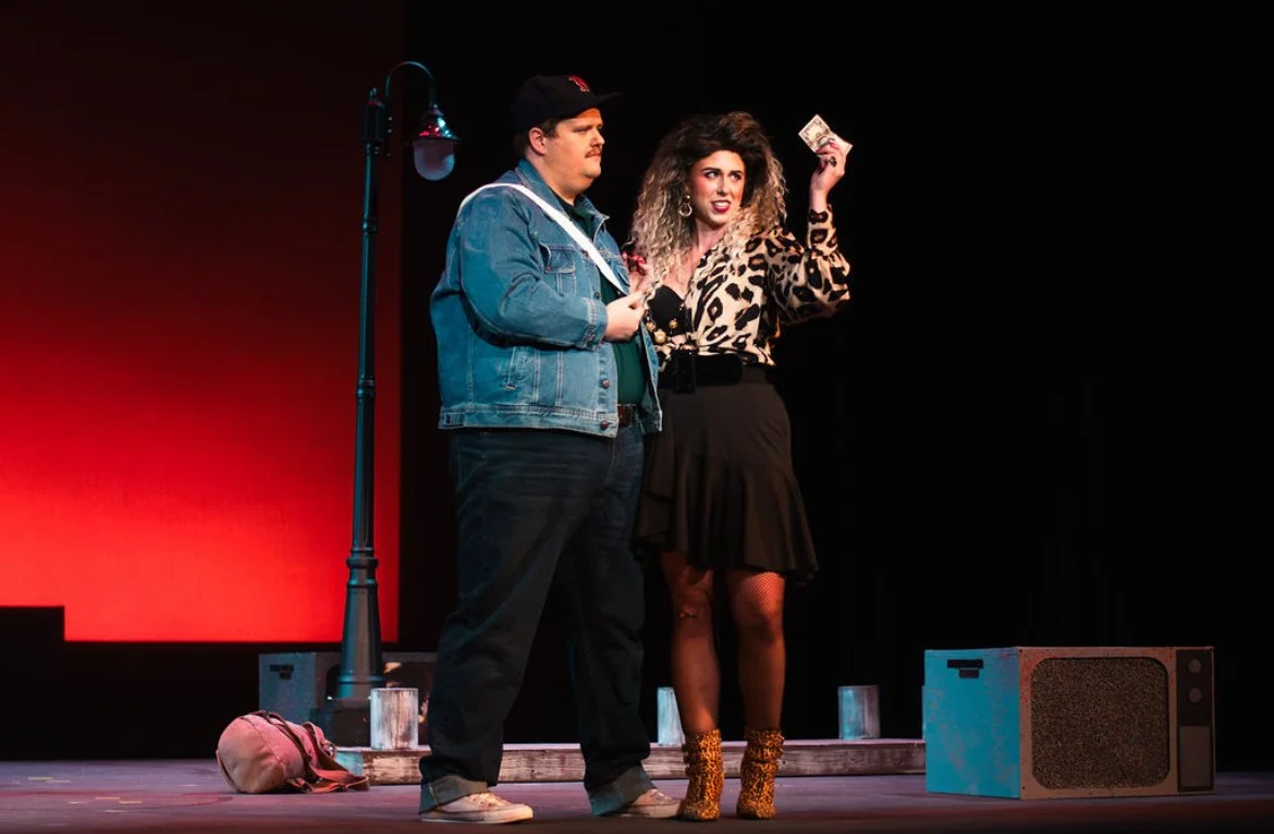 A man and woman performing on stage, with the woman holding money, in a theatrical setting.