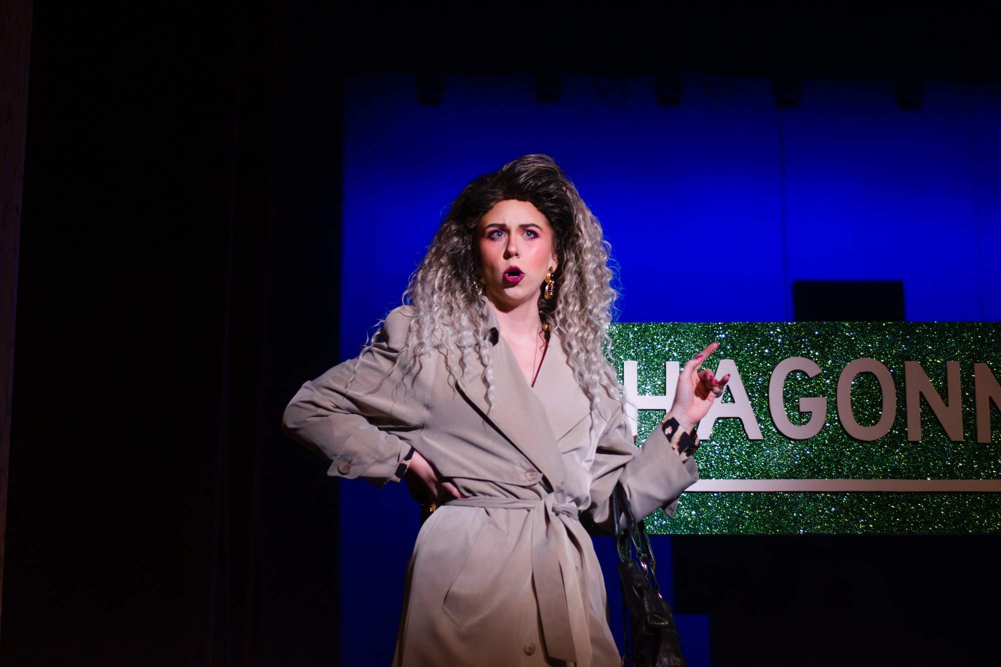 A woman with curly, blond hair, wearing a beige trench coat and large earrings, stands on stage in front of a glittery green and silver sign that reads 'HAGONN.' She has a surprised or expressive facial expression, with her right hand touching her hi