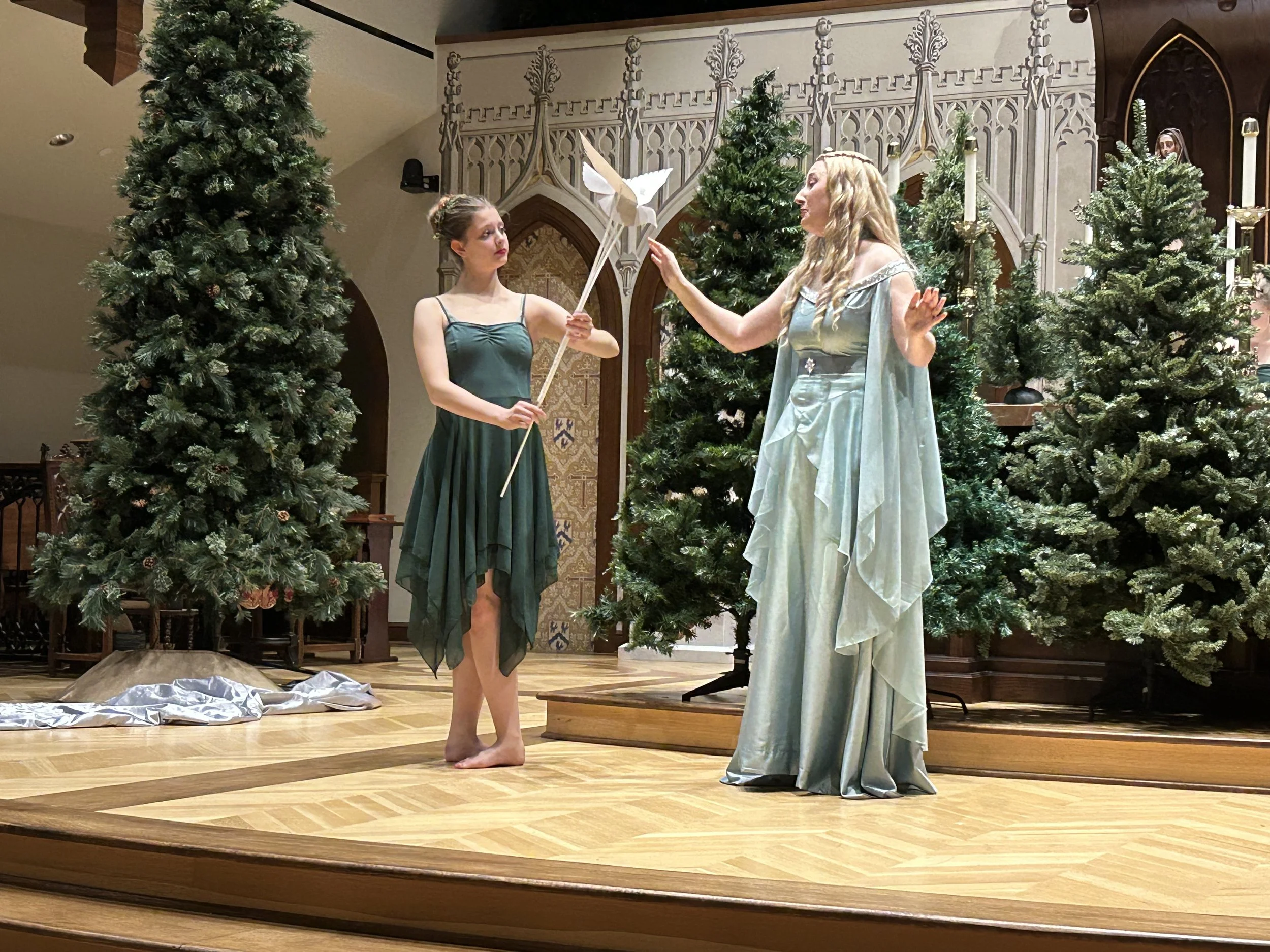 Two women dressed as fairies or mythical figures performing on a wooden stage in front of Christmas trees and church-like decor.