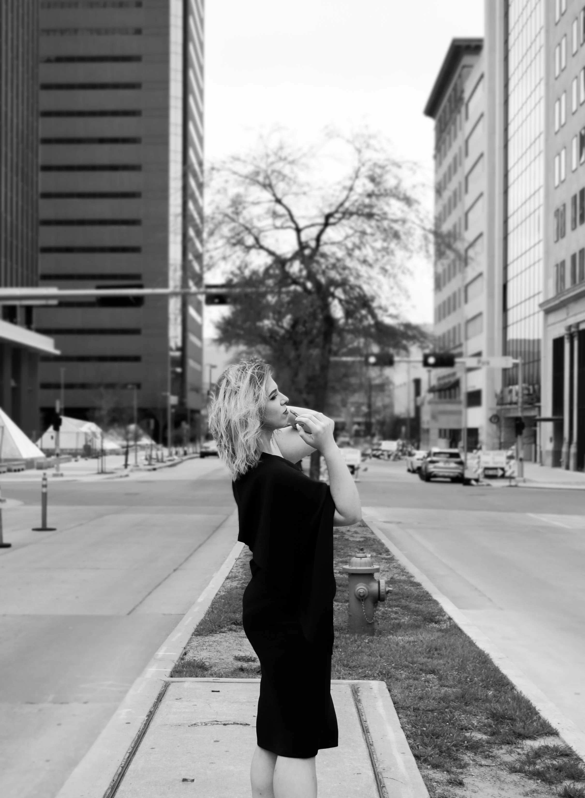 A woman with wavy blonde hair in a black dress stands on a city sidewalk, looking thoughtful with her hand near her face. The background features tall office buildings, cars on the street, and a large leafless tree, in a black and white cityscape.