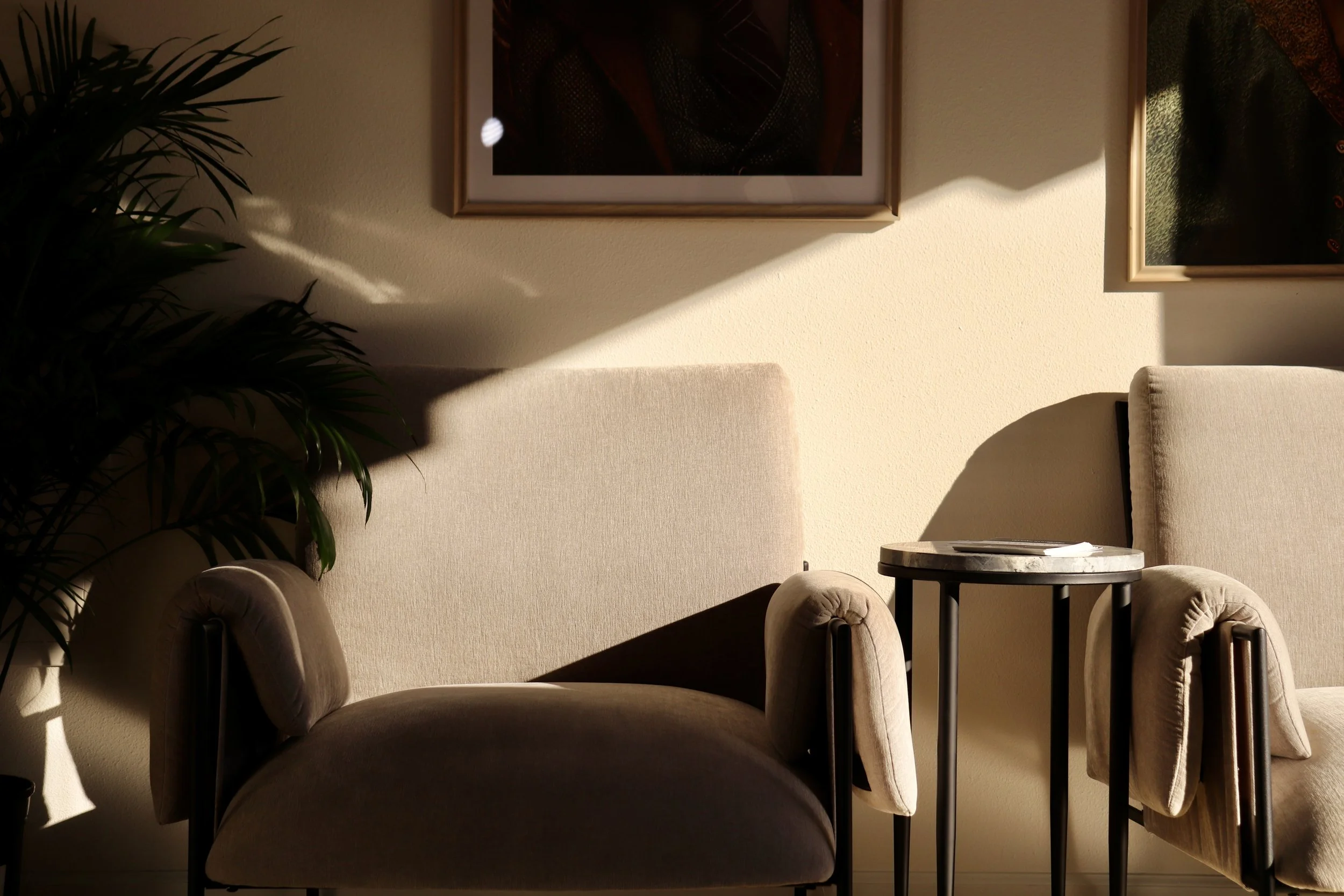 Cozy living room with beige armchairs, a small side table, wall art, and sunlight casting shadows on the wall.