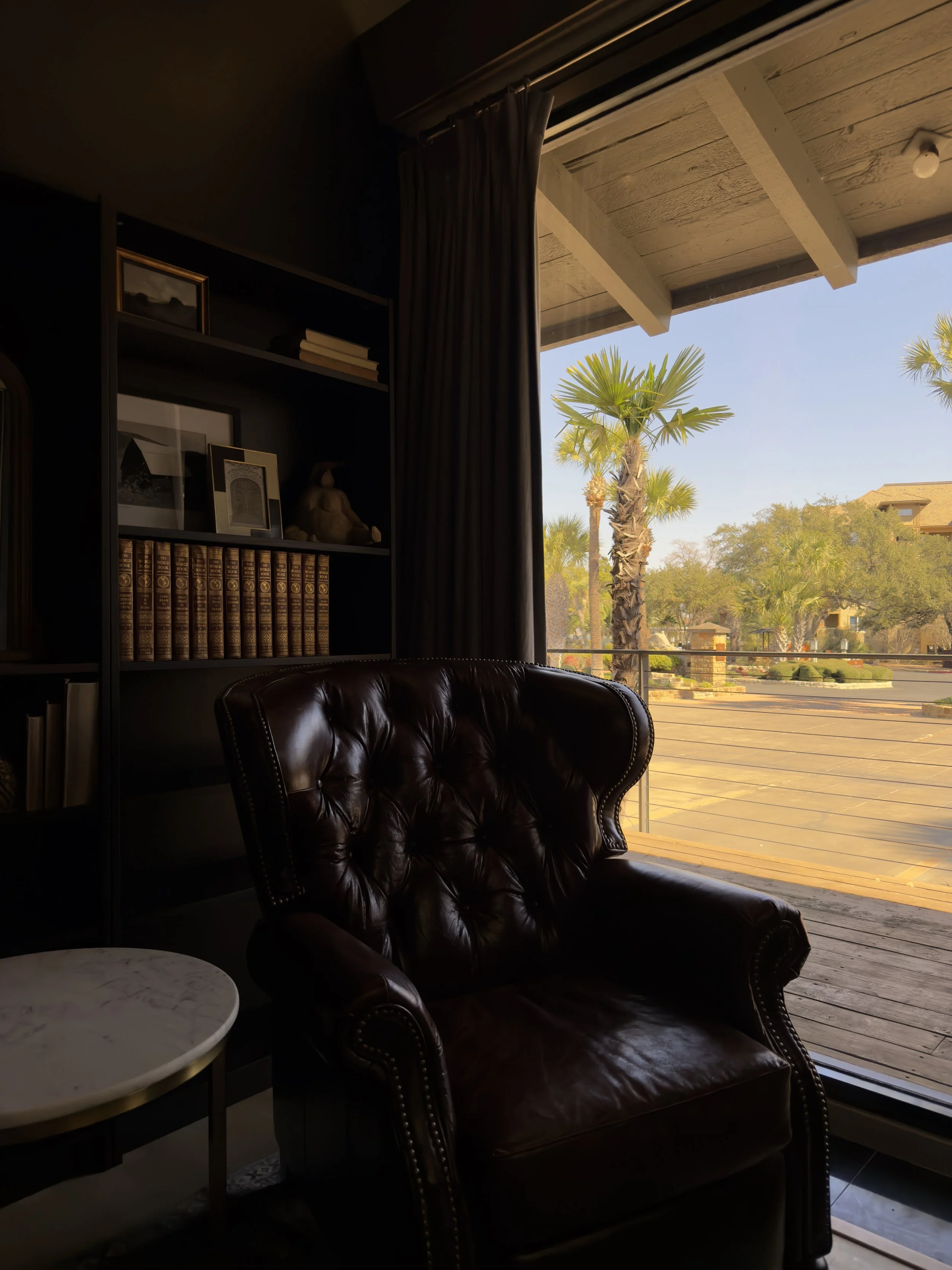 Living room with a dark leather armchair, bookshelf with books and framed art, and large open sliding door revealing a sunny outdoor patio with palm trees.