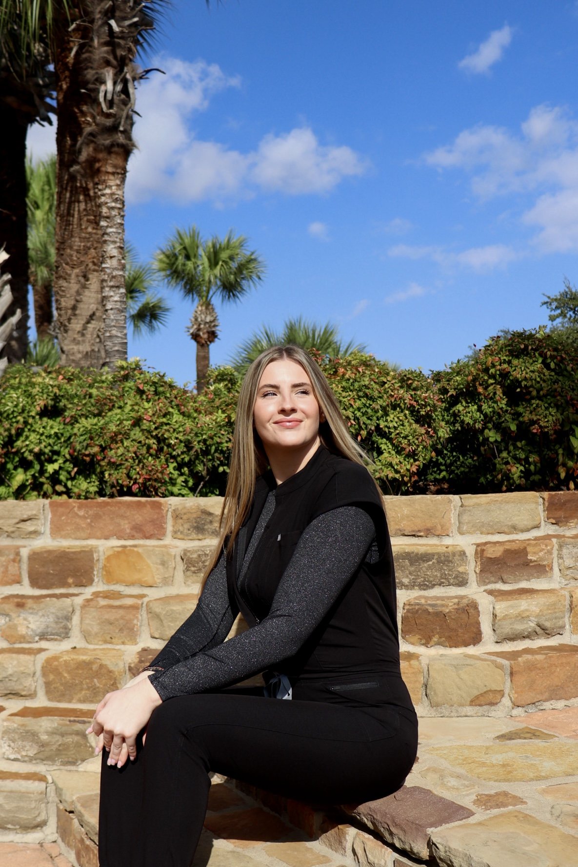A young woman with long blonde hair, wearing a black shirt, sitting on a beige sofa, smiling at the camera, with green plants and a framed poster on the wall behind her.