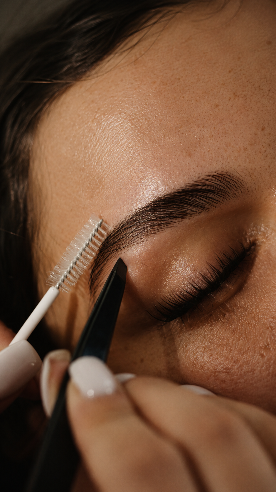 Close-up of a woman getting her eyebrows shaped with tweezers, with an eyelash brush nearby.