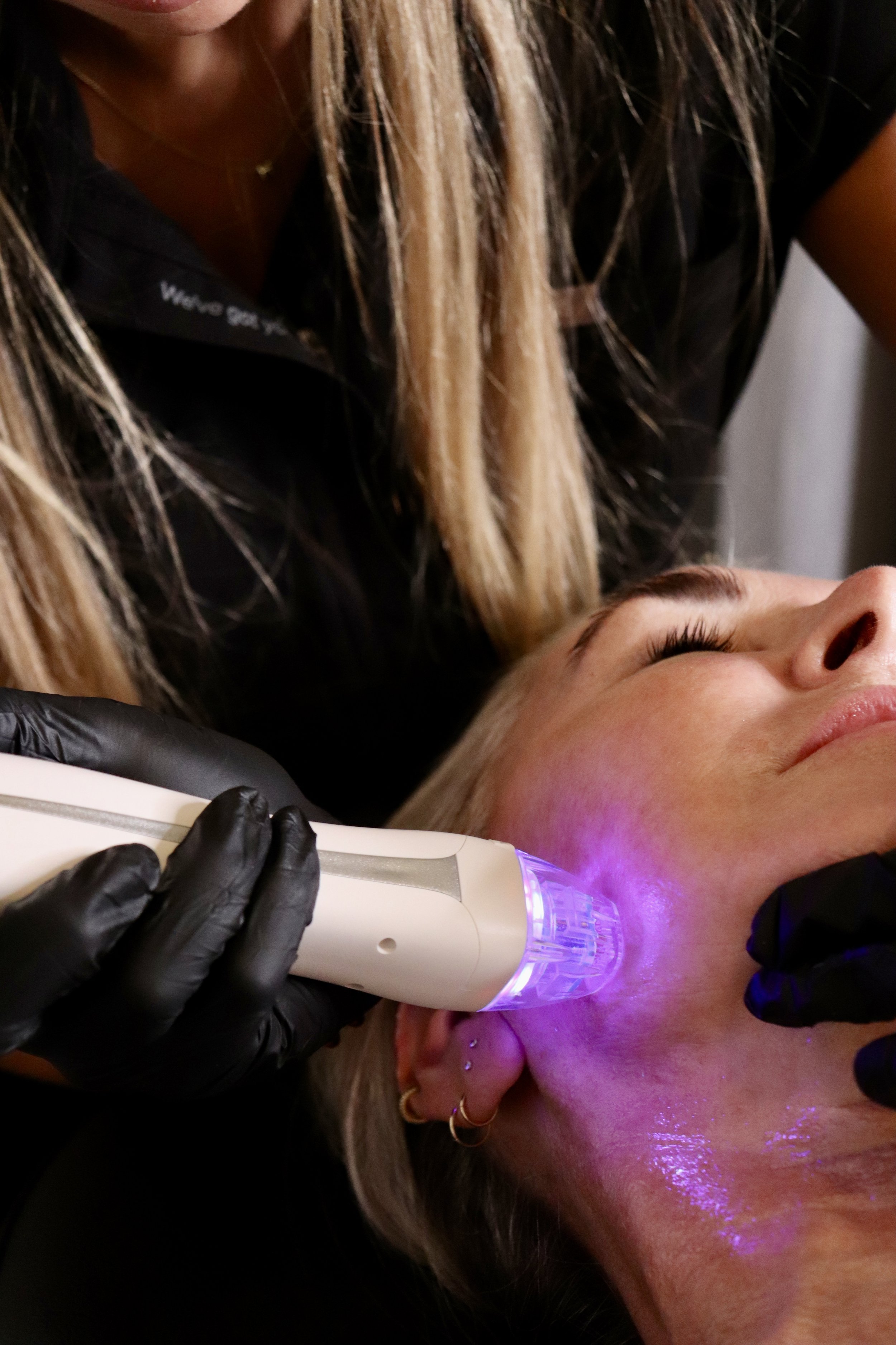 A person receives a cosmetic facial treatment with a handheld device emitting purple light, performed by a practitioner wearing black gloves.