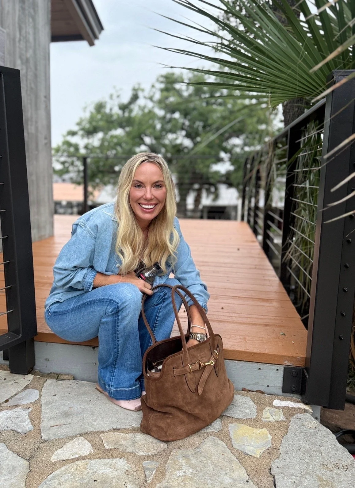 A woman with long blonde hair, wearing a denim jacket and jeans, crouches on a wooden deck holding a water bottle. She smiles at the camera with a brown handbag on the ground beside her, and plants and trees are visible in the background.