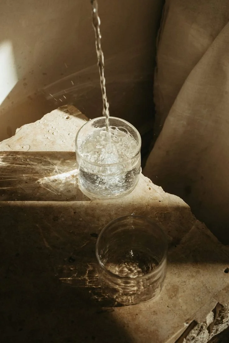 A glass being filled with water, with another empty glass below on a rough stone surface. Sunlight is casting shadows on the surface.