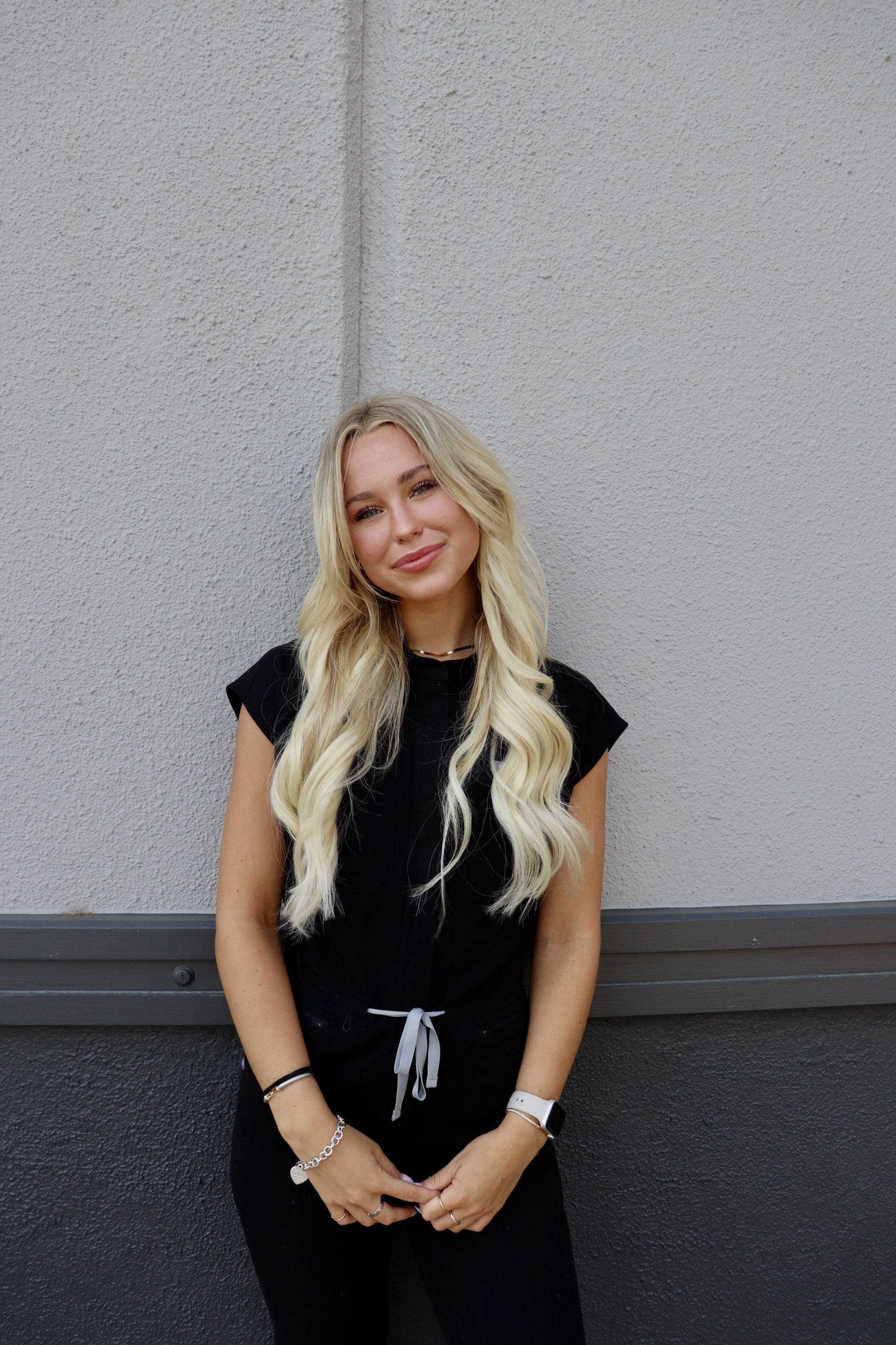 A young blonde woman with long wavy hair standing against a textured gray wall, wearing a black sleeveless top, black pants, and accessorized with bracelets, a watch, and a necklace.