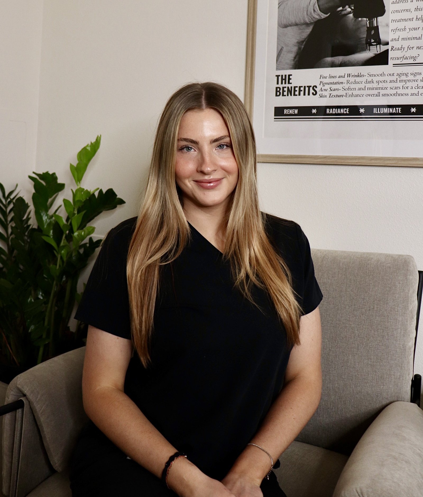 A young woman with long blonde hair, wearing a black shirt, sitting on a beige sofa, smiling at the camera, with green plants and a framed poster on the wall behind her.