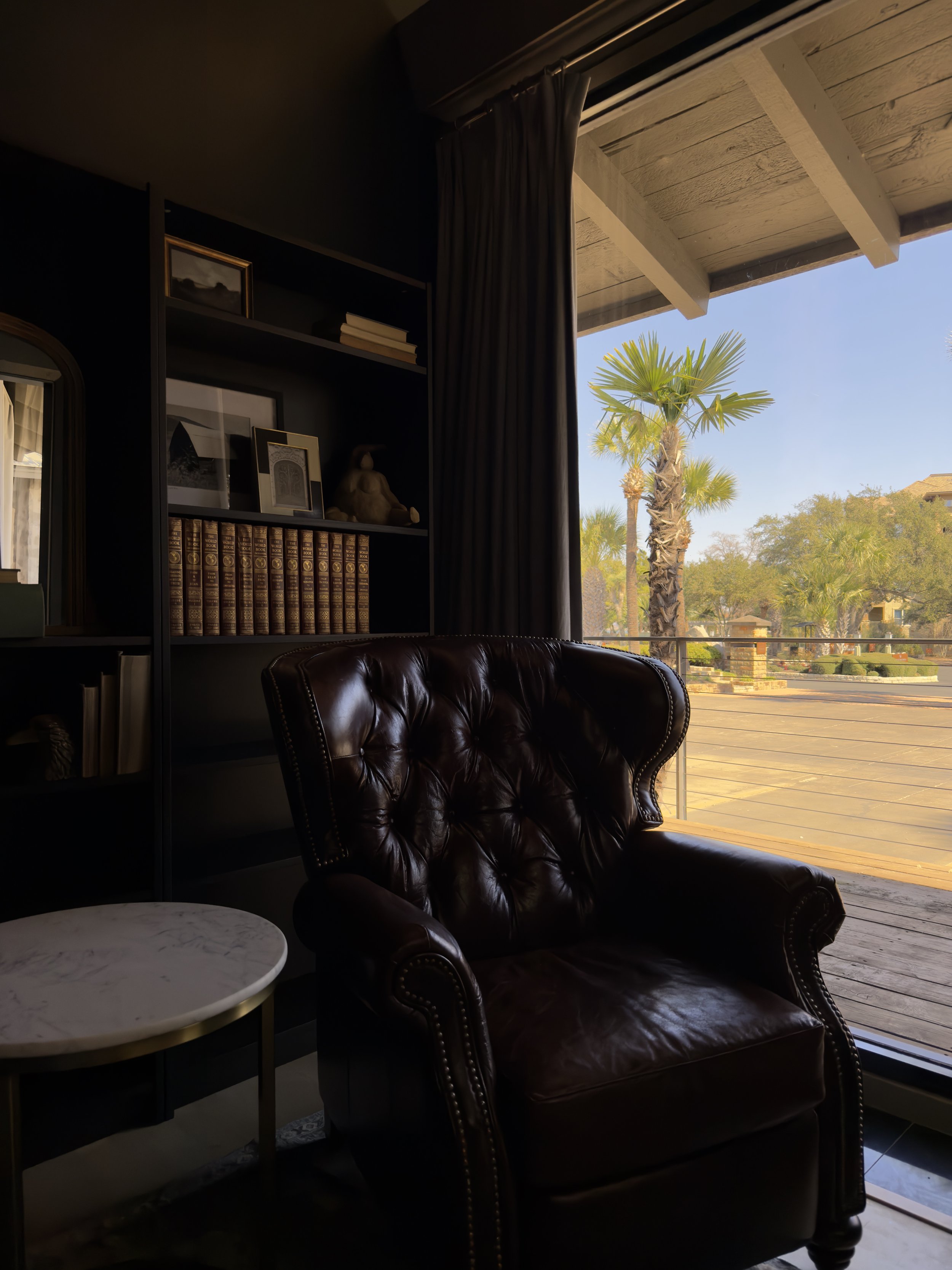 Interior living room with a leather armchair, bookshelf with books and decor, large sliding glass door with curtains, view of palm trees and a sunny outdoor landscape.