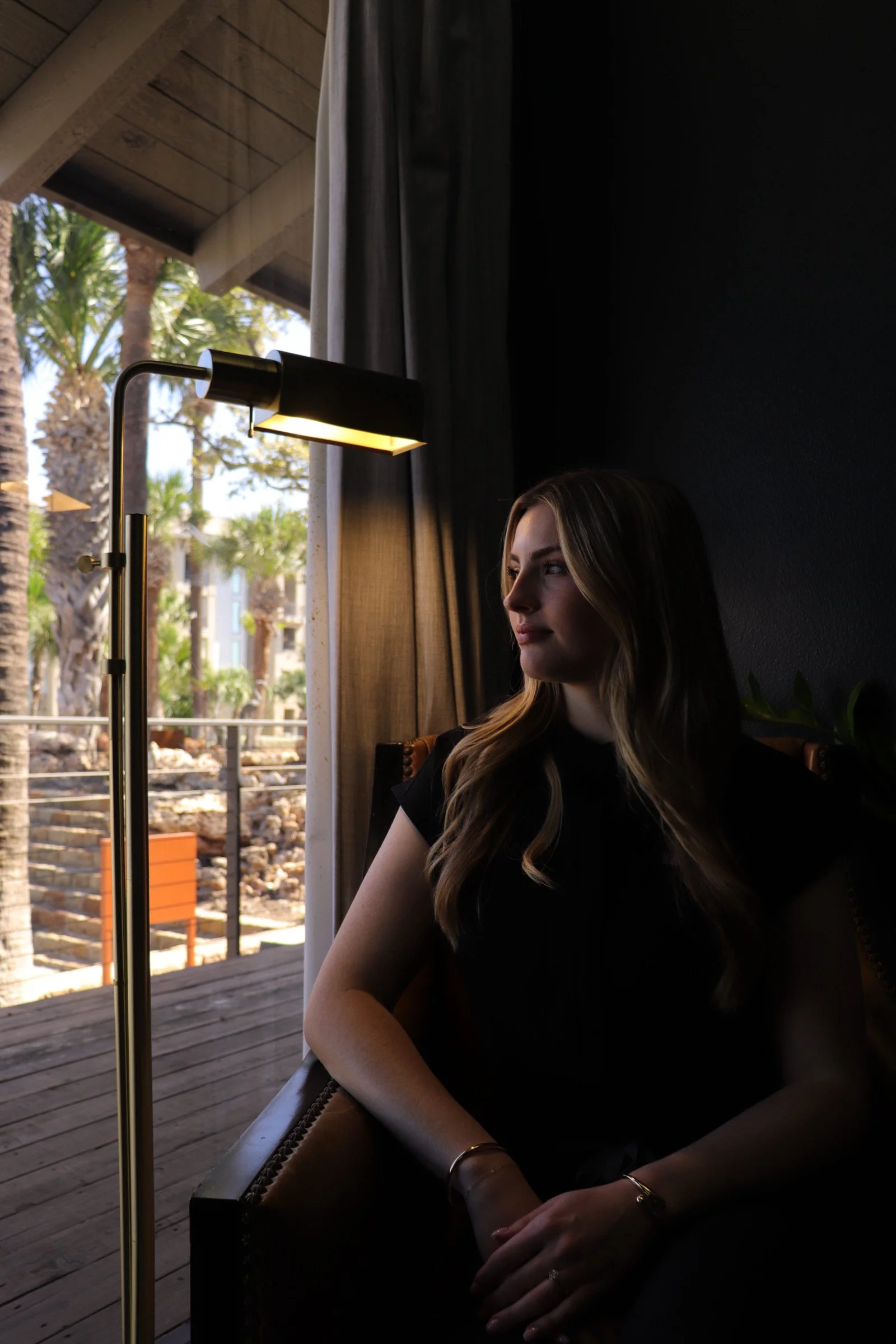 A woman with long wavy hair sitting indoors beside a window, looking outside with natural light illuminating her face, and tropical trees visible outside.