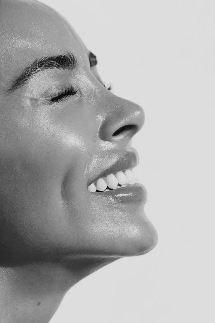 A close-up black and white photo of a smiling woman with her eyes closed, showing her teeth.