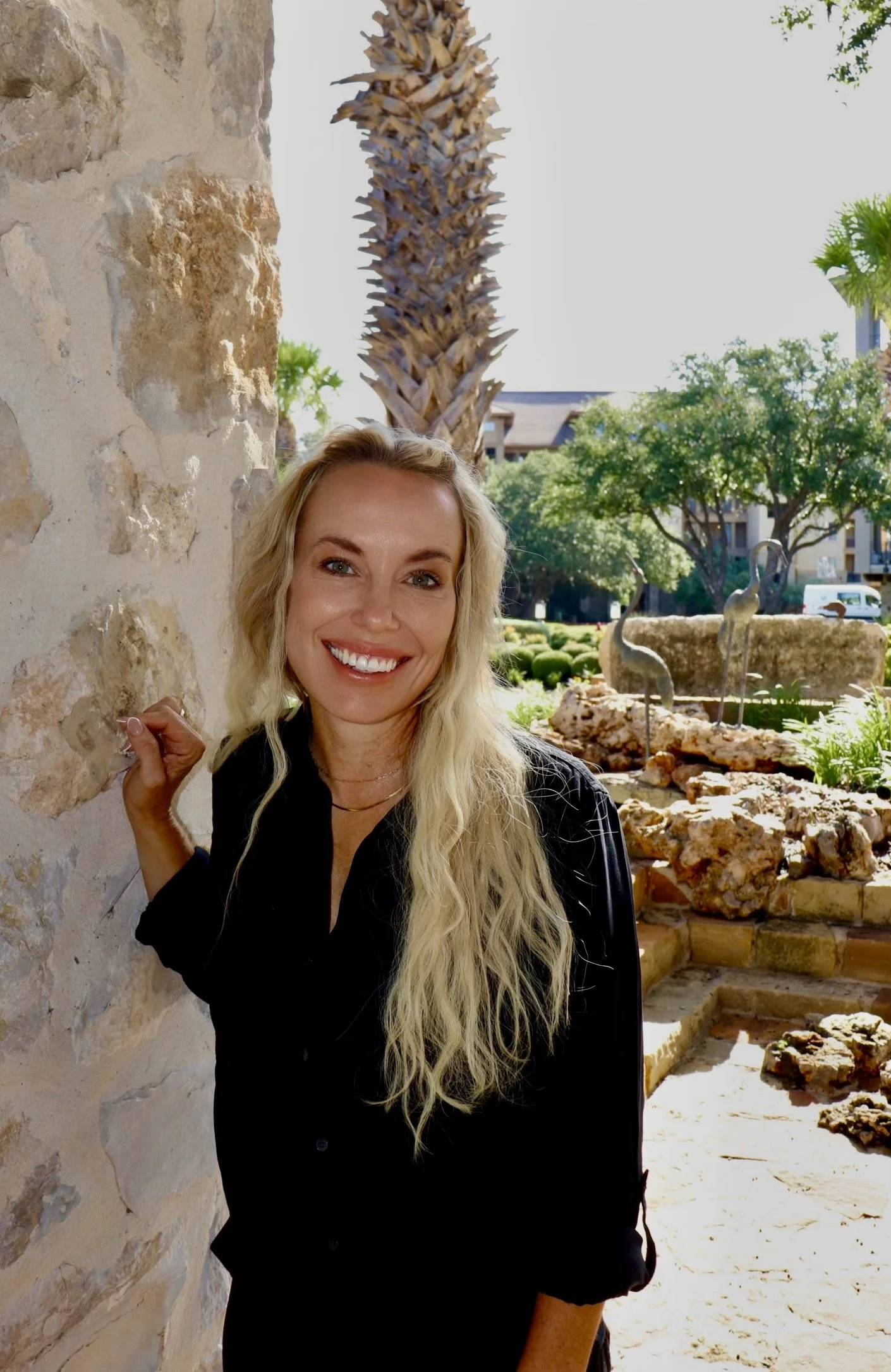 A woman with long blonde curly hair and a black shirt smiling while standing beside a stone wall outdoors. Behind her, there is a landscaped garden with a palm tree, decorative bird sculptures, and other trees, with buildings and a vehicle visible in the background.