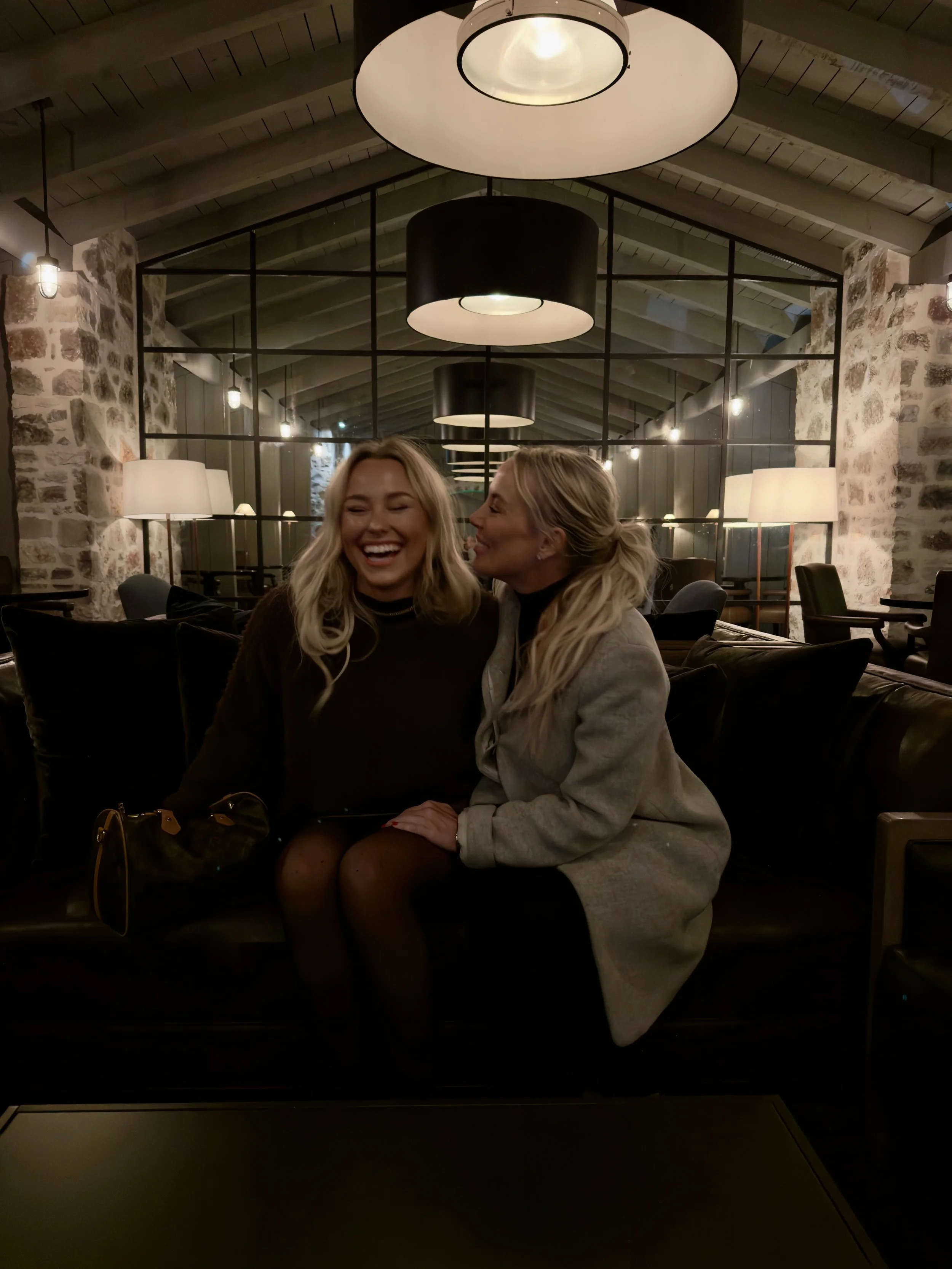 Two women sitting on a dark leather couch in a dimly lit, modern lounge with exposed stone walls, large glass mirror, and hanging ceiling lights, sharing a laugh.