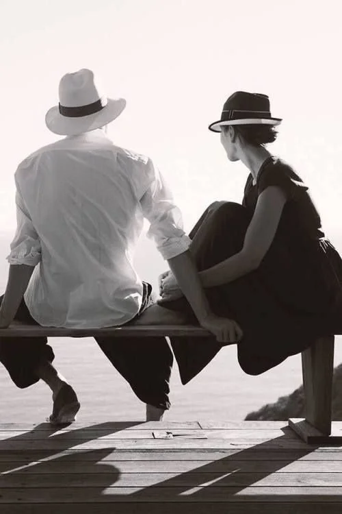 Two people sitting on a bench by the water, wearing hats and casual clothing, engaging in conversation.
