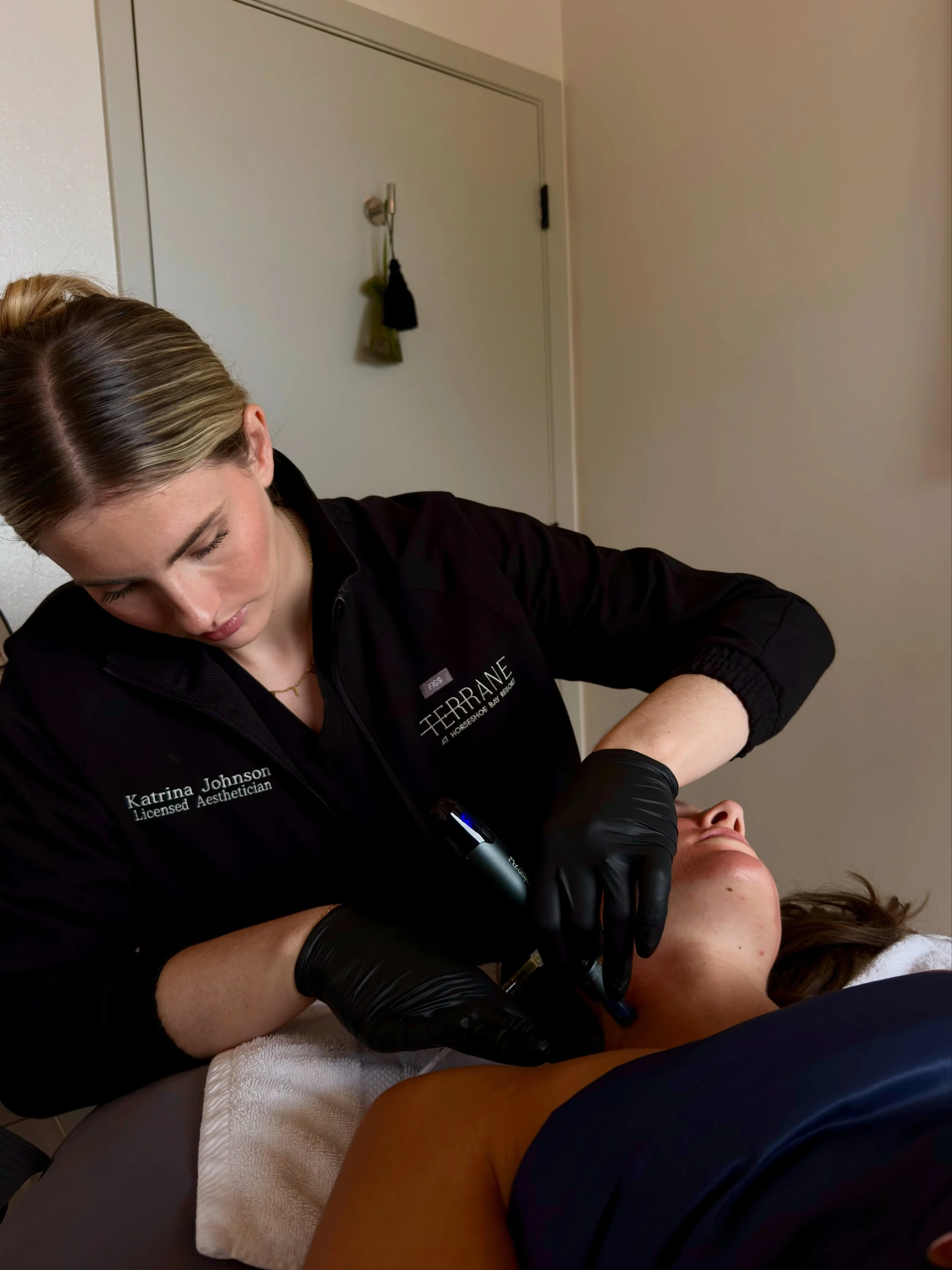 A woman receiving a facial treatment from an aesthetician in a Spa setting, with her lying on a treatment bed, and the aesthetician wearing black gloves and a black uniform.