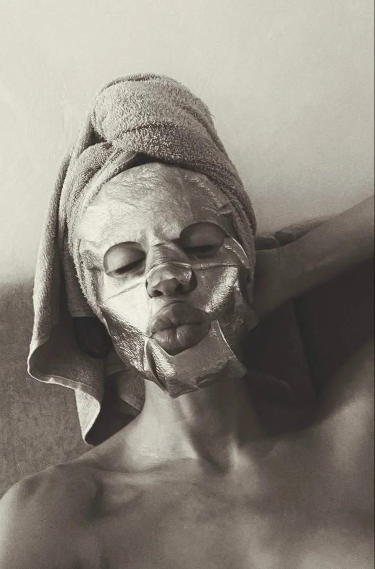 A person with a towel wrapped around their head, wearing a silver facial mask and facial peel strips, relaxing with eyes closed.