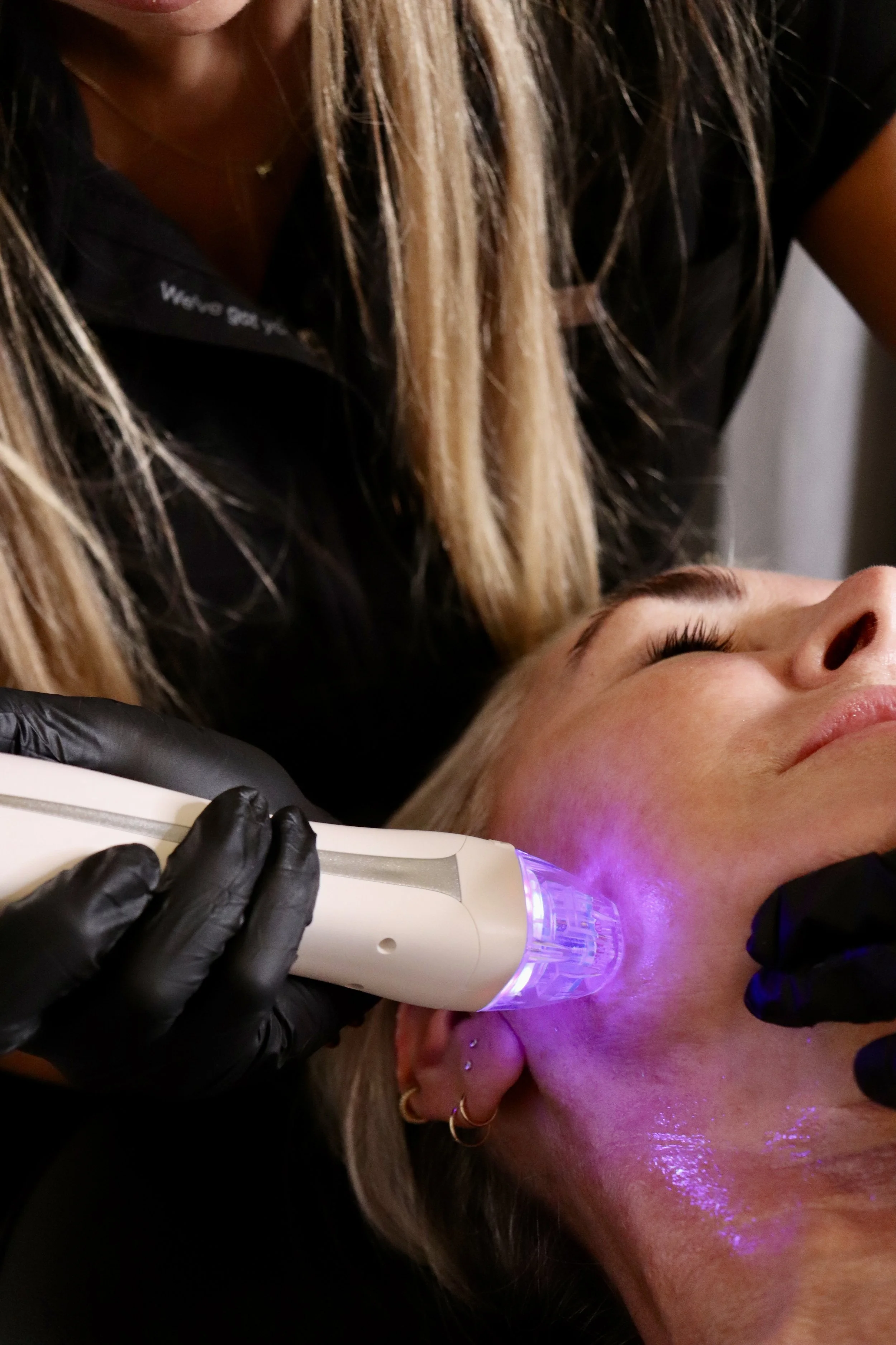 A woman receives a facial treatment with a handheld device emitting purple light, while lying down with her eyes closed, and a practitioner dressed in black gloves holds the device near her face.