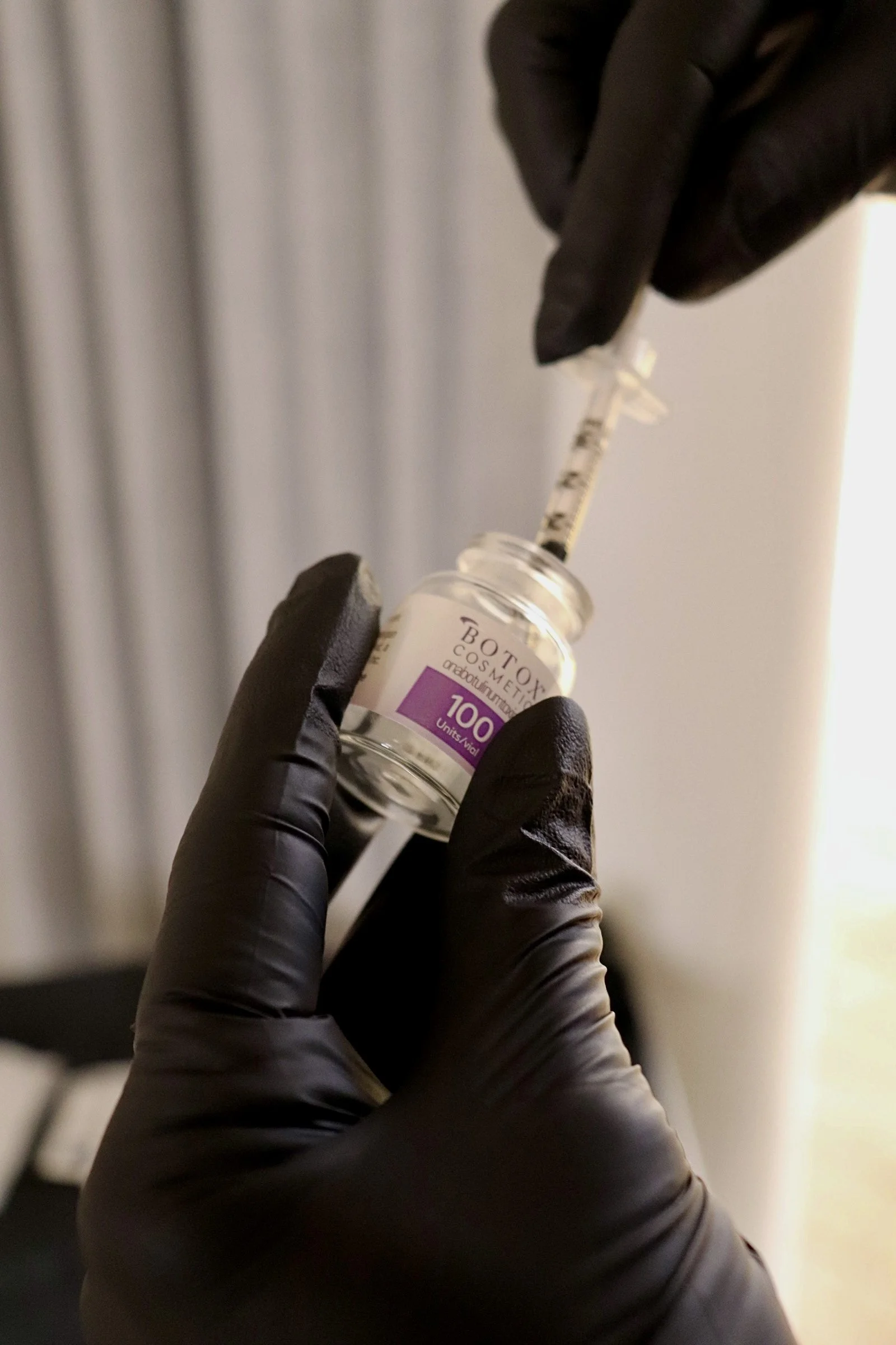 A person wearing black gloves drawing a clear liquid from a small vial labeled "Botox Cosmetic" with a syringe.