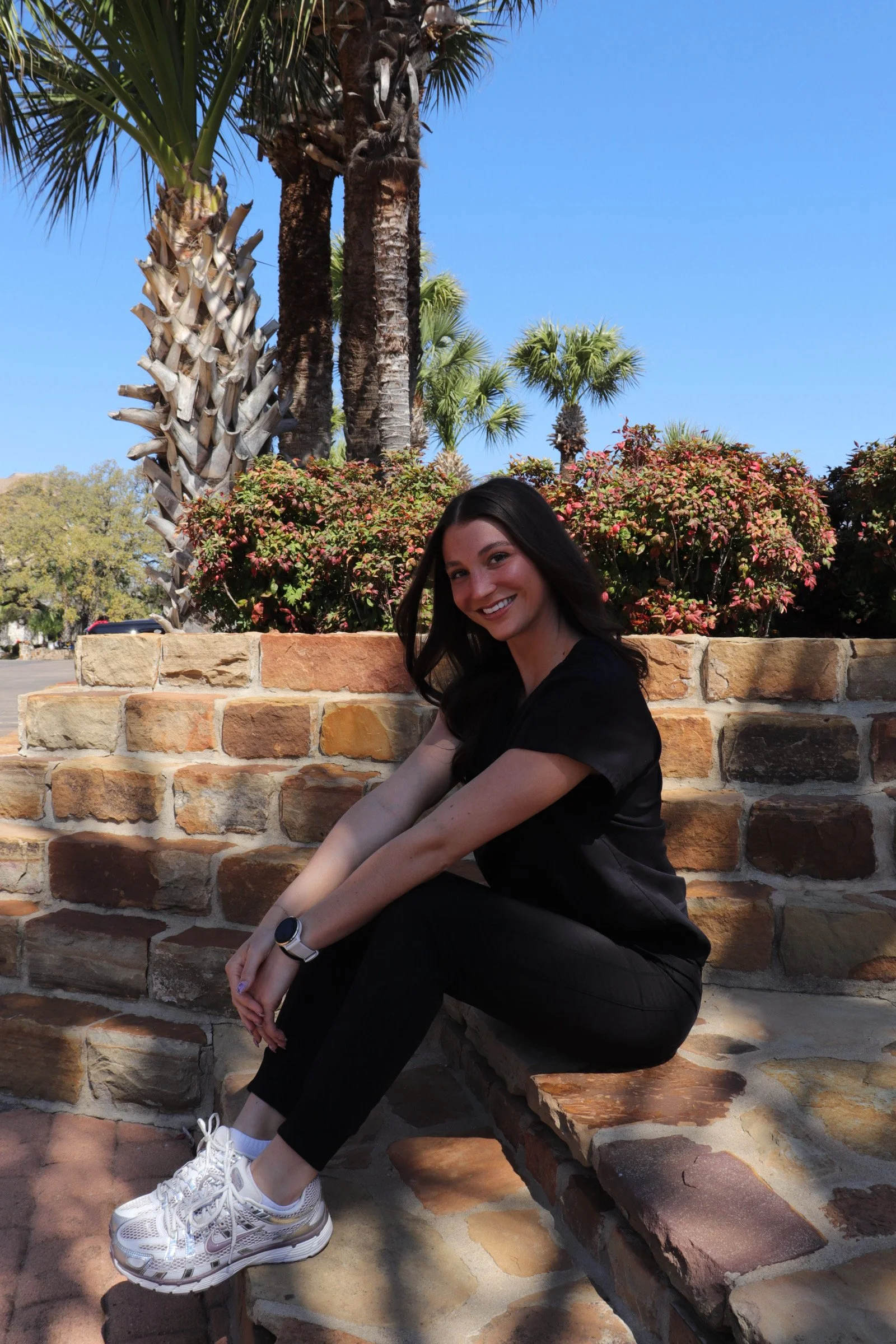 A woman with long dark hair wearing a black top and black pants sitting on a brick ledge outdoors. Behind her are palm trees, shrubs, and a clear blue sky. She is smiling, with her arms resting on her knees, and is wearing a smartwatch on her left wrist and white sneakers with gray details.