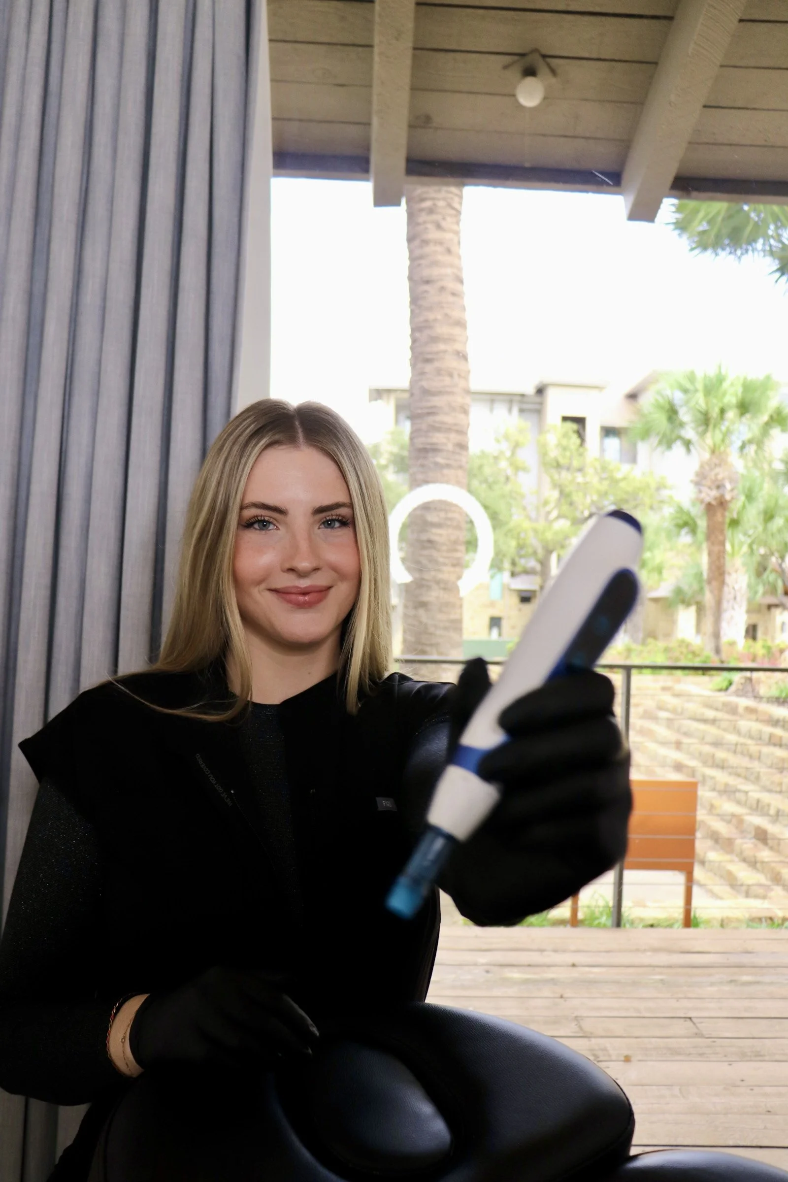 A woman with blonde hair taking a selfie with a smartphone, sitting outdoors with palm trees and buildings in the background.