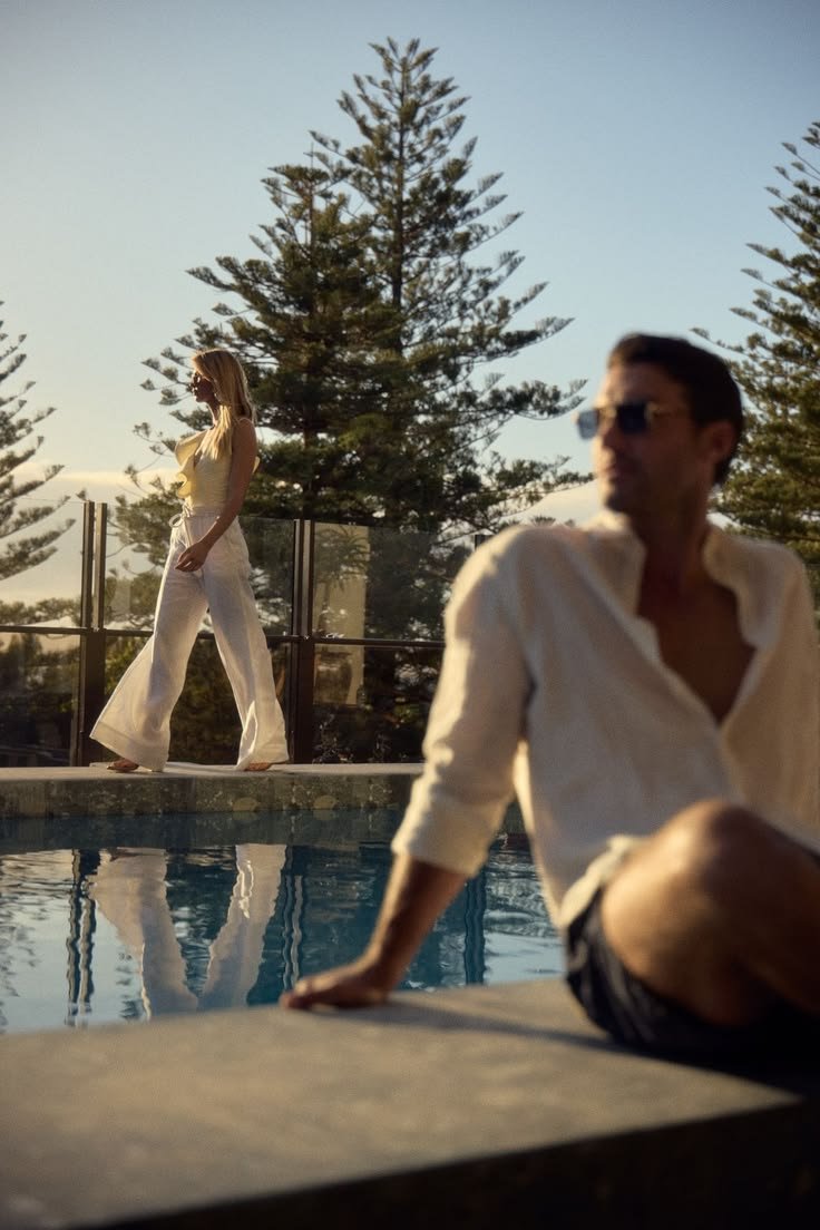 A woman walking along the edge of a swimming pool at sunset, wearing white pants and a yellow top, with tall trees in the background. A man is sitting nearby by the pool, wearing sunglasses and a light-colored shirt, with his legs crossed.