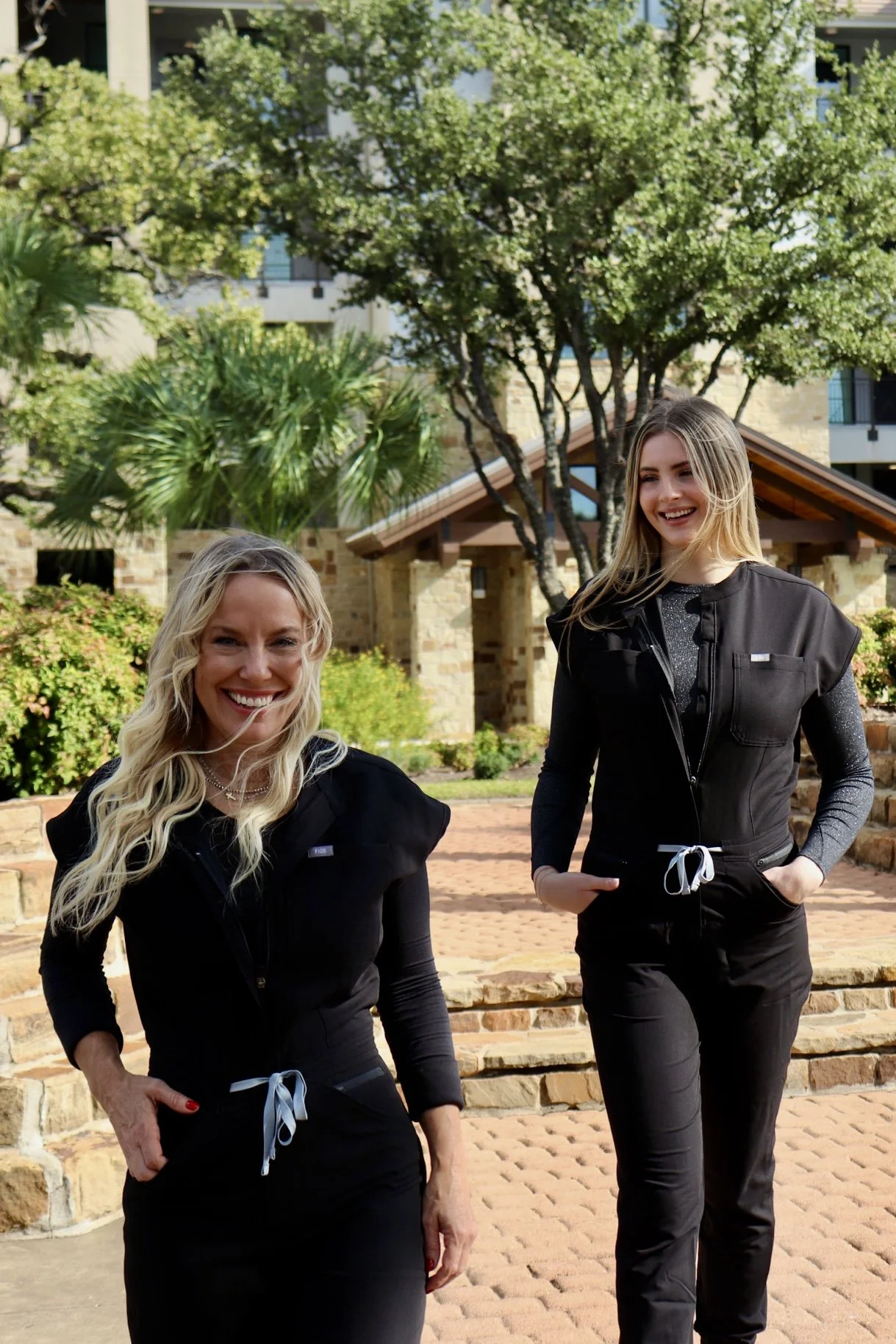 Two smiling women in black outfits standing outdoors in front of a house with trees and greenery.