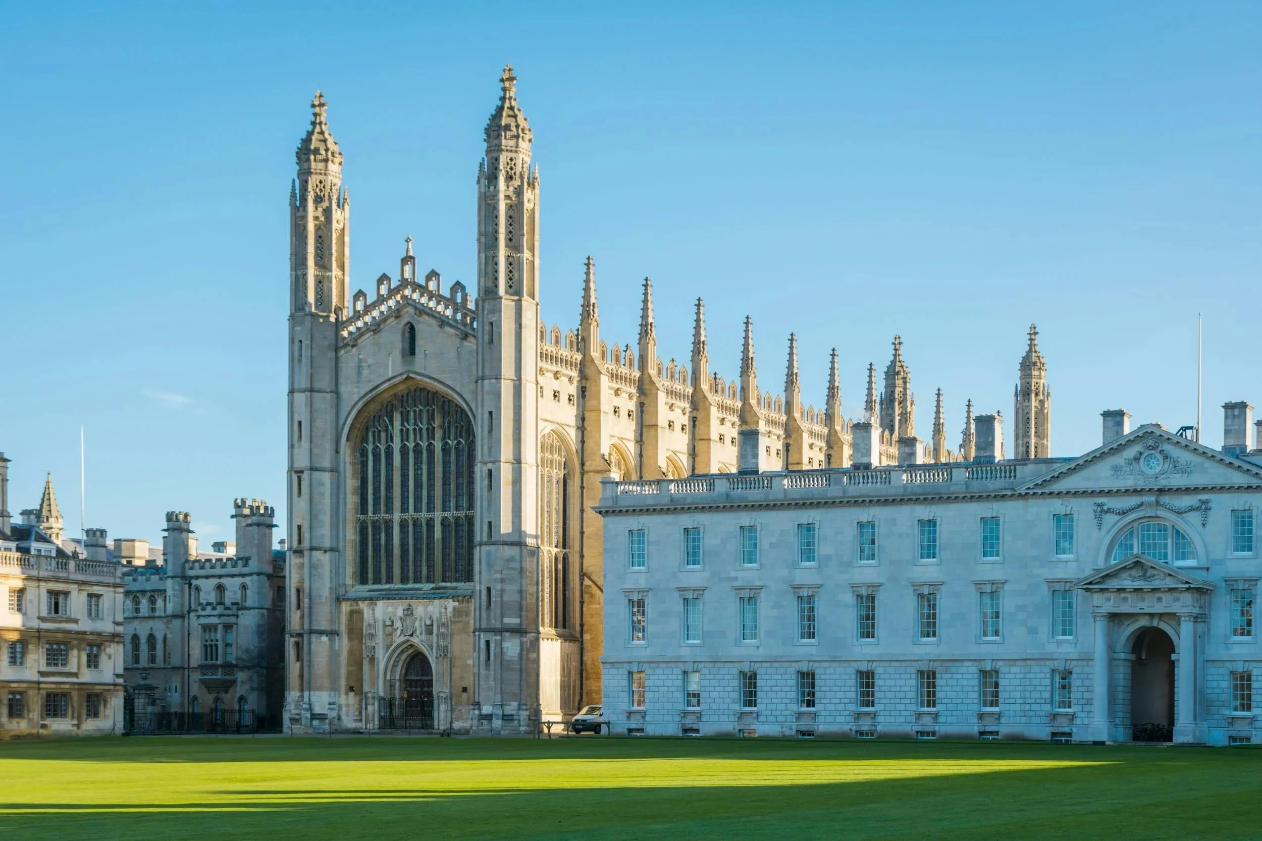 A large historic cathedral with Gothic architecture, tall spires, and intricate stone detailing, set against a clear blue sky with green grass in the foreground.