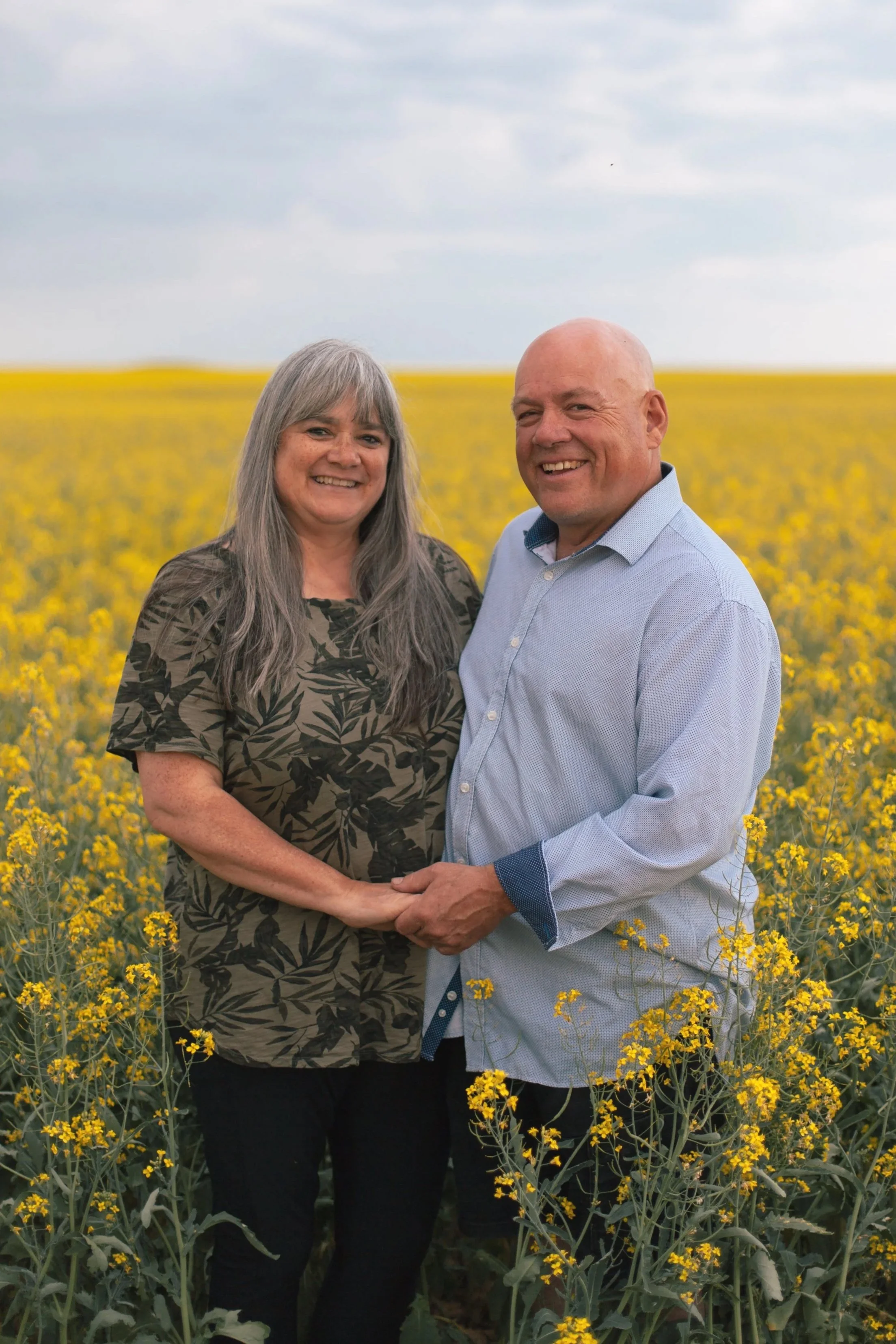 A smiling woman and a smiling man holding hands in a yellow flower field under a cloudy sky.