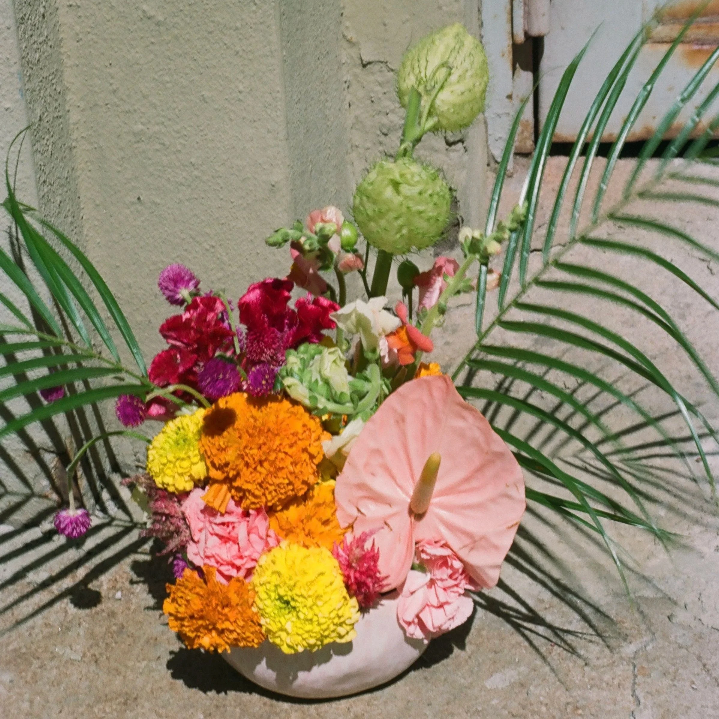 Colorful flower arrangement with pink anthurium, marigolds, and other various flowers, accented by green palm leaves in a pink bowl, against a beige wall background.