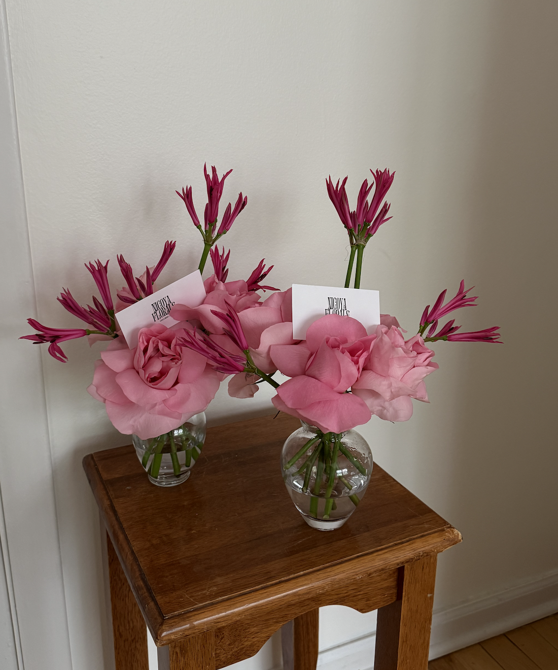 Two glass vases with pink flowers, including large pink roses and smaller pink and purple flowers, placed on a wooden table against a plain wall.