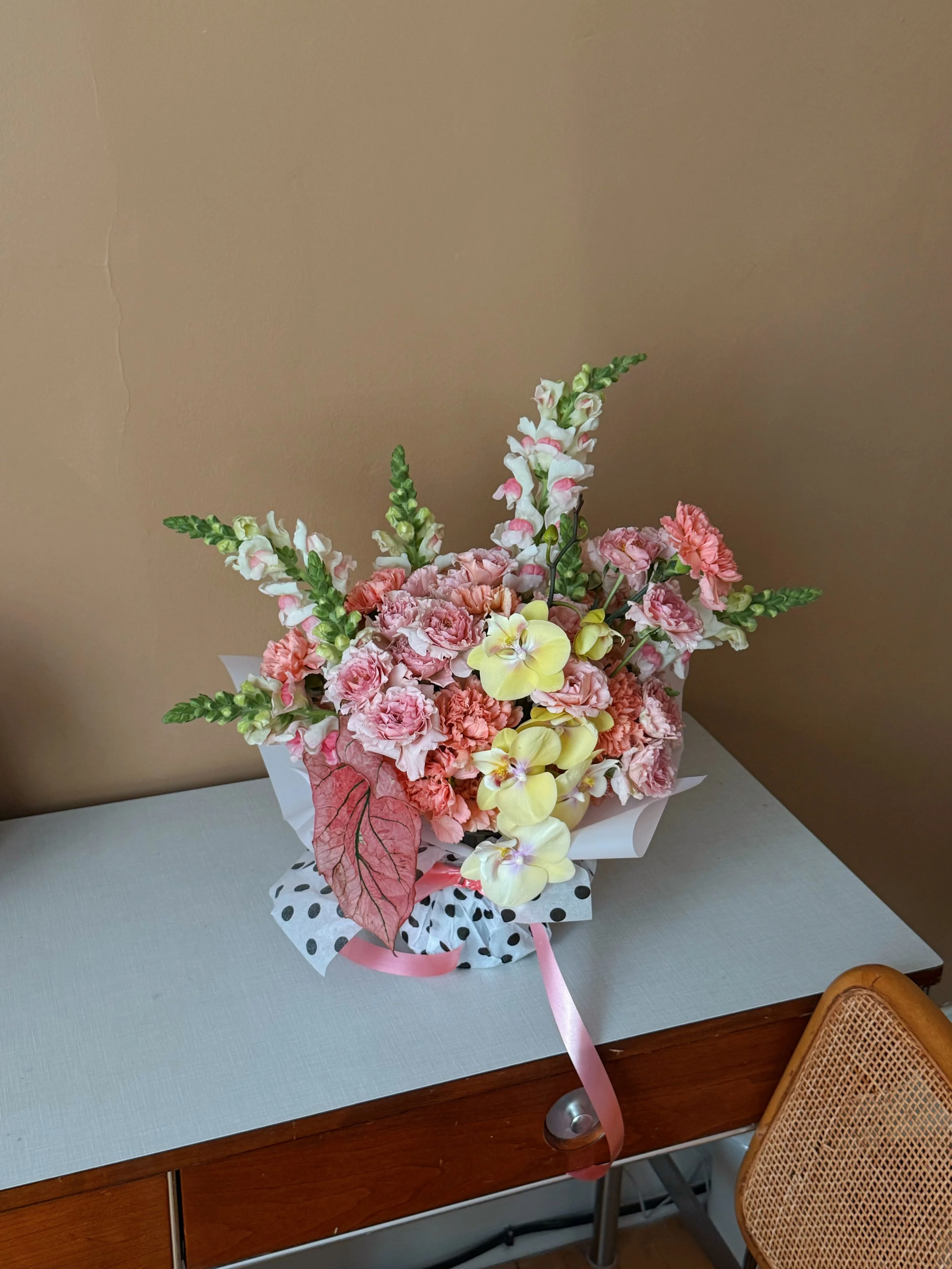 A bouquet of pink, yellow, and white flowers, including orchids and snapdragons, arranged with pink and red leaves in a decorative wrapping with a pink ribbon, placed on a gray surface against a brown wall.