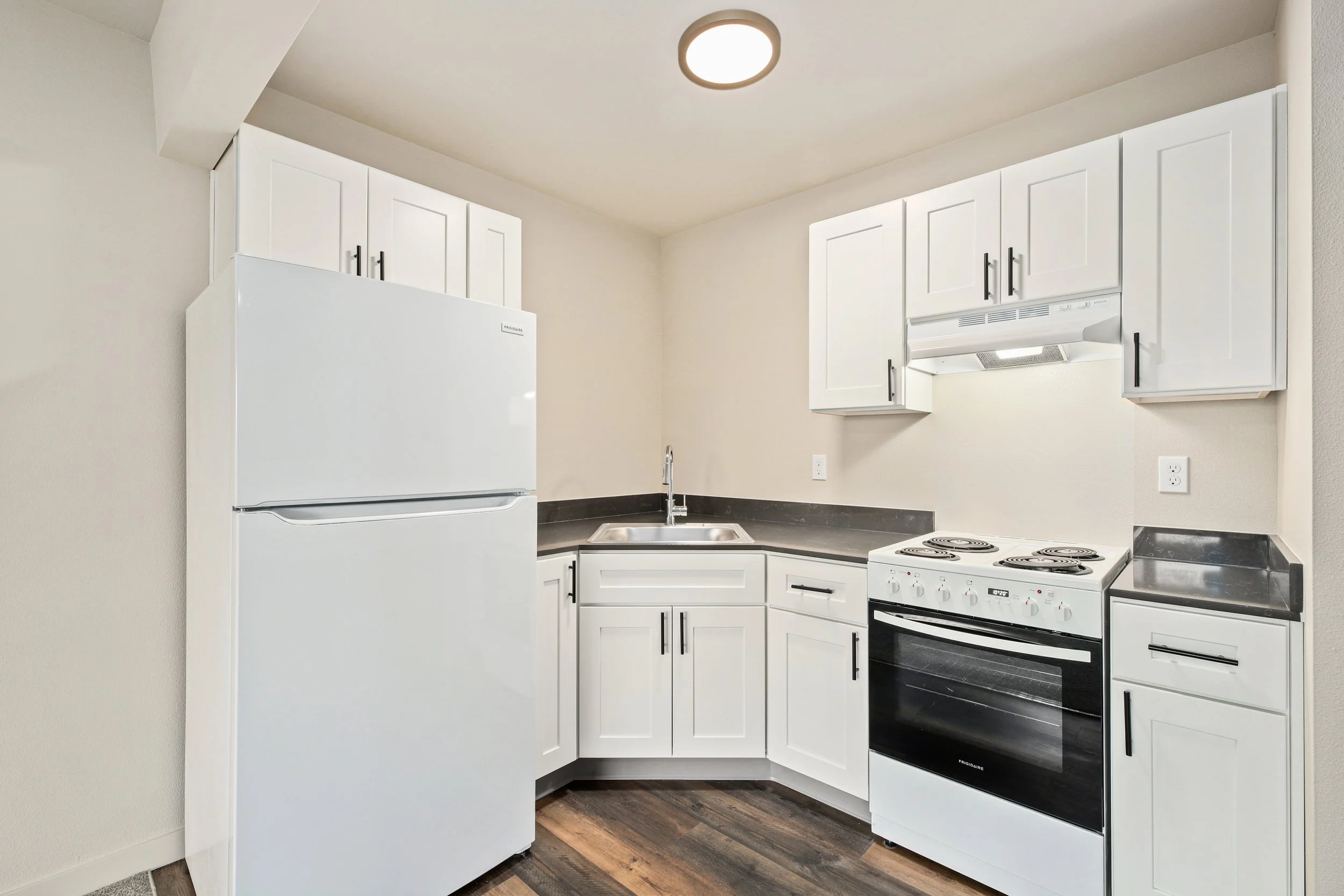 A small kitchen with white cabinets, a white refrigerator, a white stove with four burners, a stainless steel sink, and dark countertops. The floor is wood, and there's a ceiling light fixture.