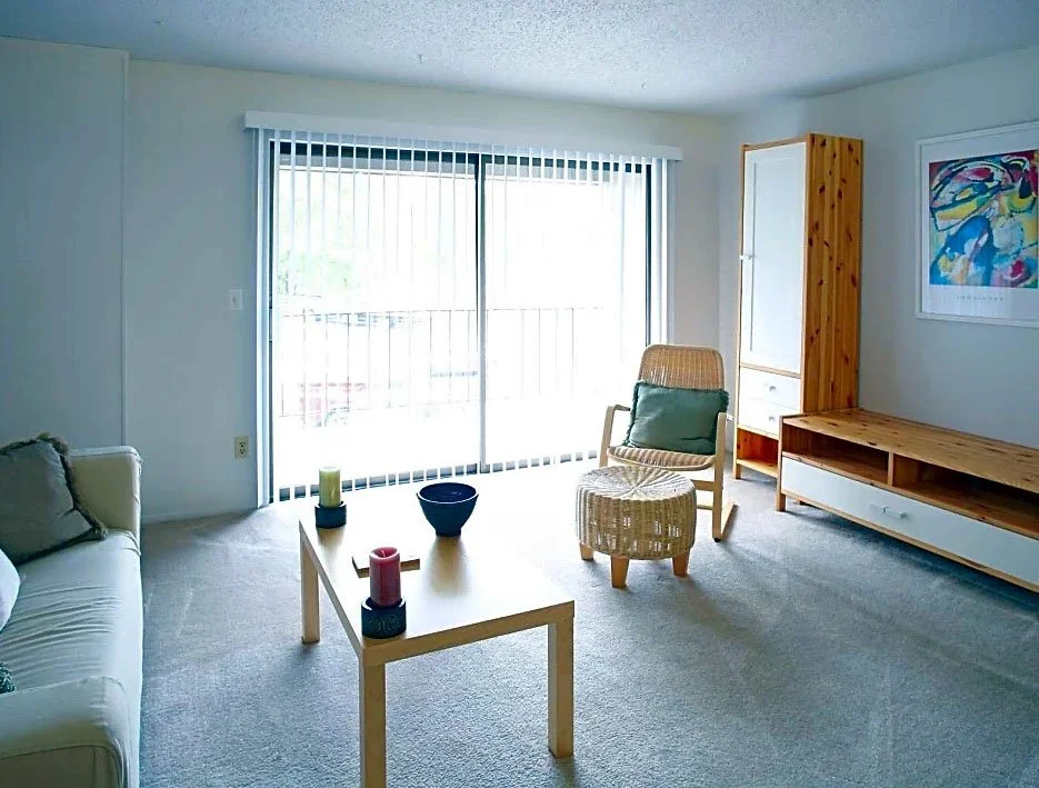 Living room with sliding glass door, light-colored sofa, small wooden table with candles, rattan chair with cushion, wooden cabinet, and colorful artwork on the wall.