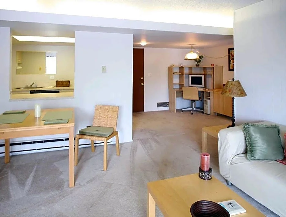 Living room and dining area in a home with light-colored walls and carpet, a small dining table with chairs, a couch with pillows, a desk with a computer, and a kitchen view in the background.