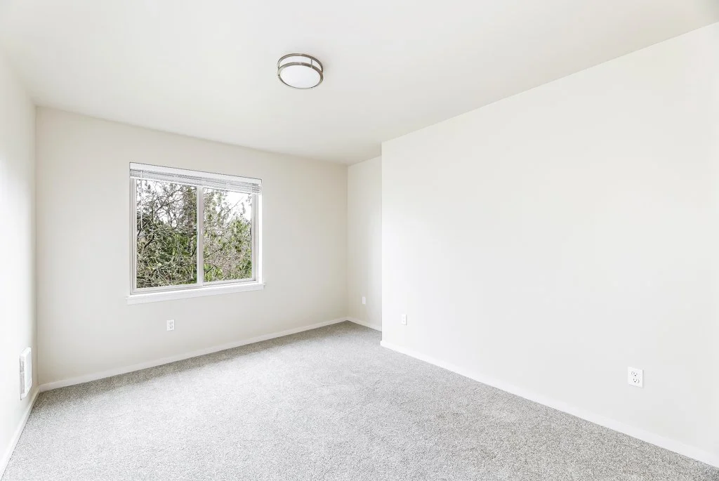 Empty room with white walls, a window showing trees outside, gray carpet, ceiling light, and electrical outlets.