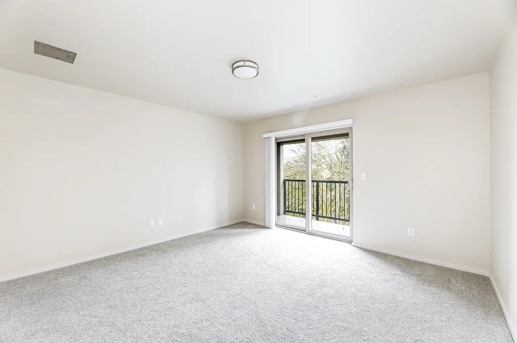 Empty living room with sliding glass door leading to a balcony, beige carpet, white walls, and ceiling light fixture.
