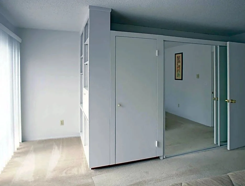 A room with white walls, beige carpet, and a built-in white shelving unit. There is a sliding glass door with vertical blinds on the left. A small framed picture hangs on the wall in the adjacent room, which has an open doorway with gold doorknobs.