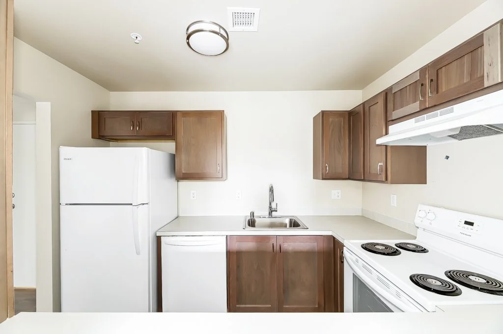 Kitchen with white refrigerator, white stove, stainless steel sink, brown wooden cabinets, and white walls.