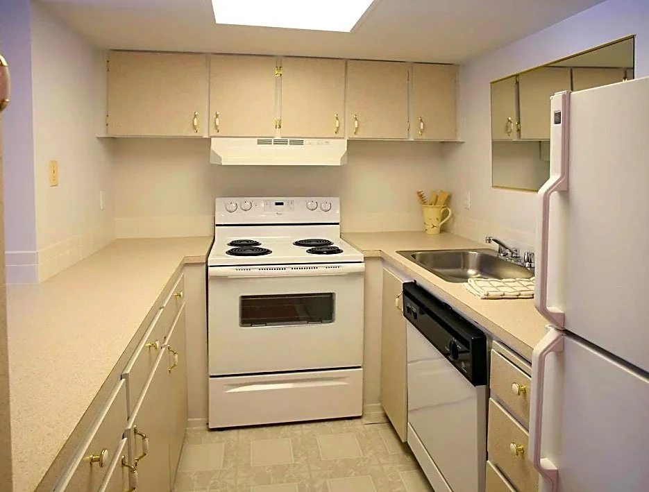 A compact kitchen with beige cabinets, white appliances including a refrigerator, oven, and microwave, a stainless steel sink, and a light countertop.
