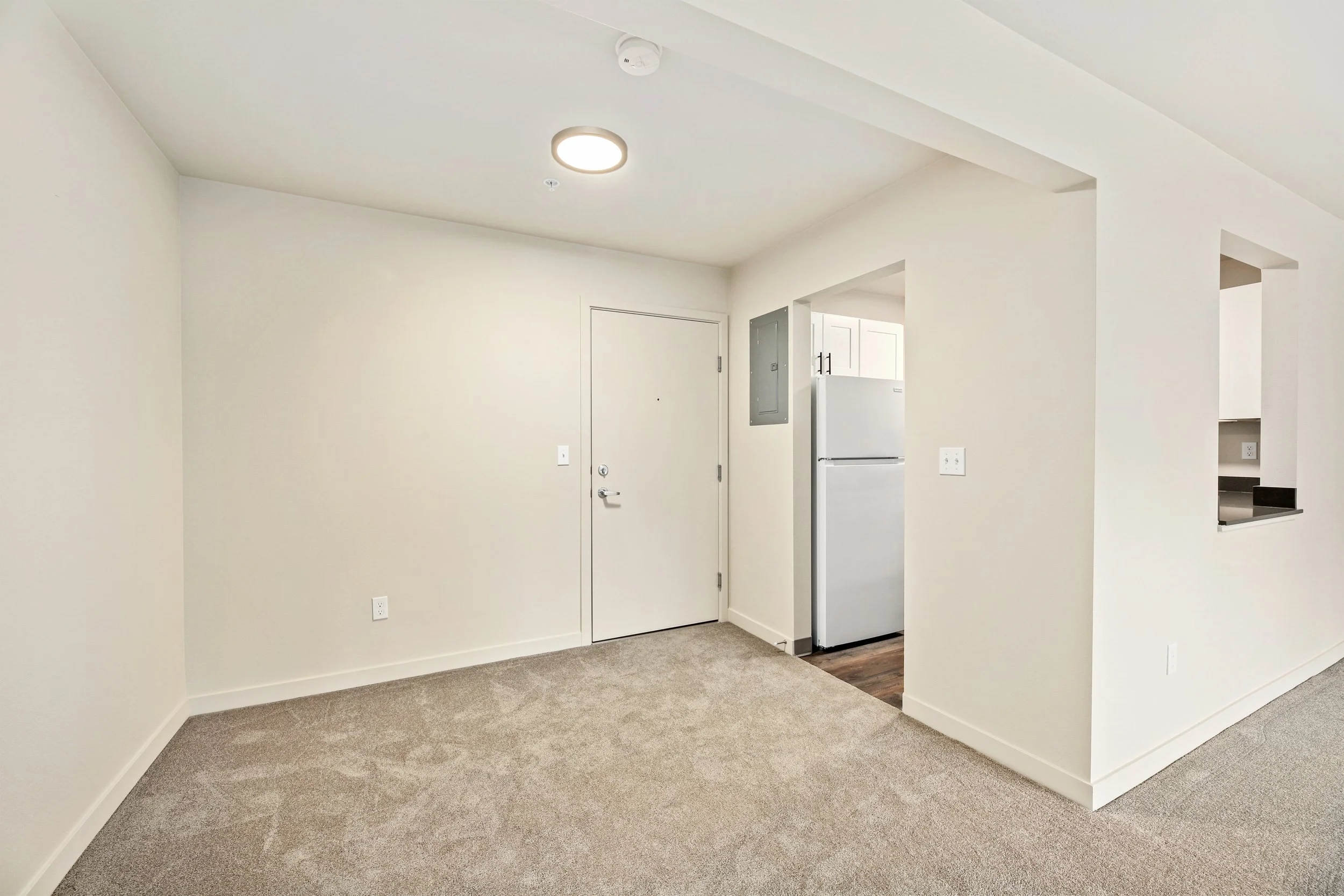 Empty living room with beige carpet, white walls, and a ceiling light, adjacent to a kitchen area with a partial view of a white refrigerator.