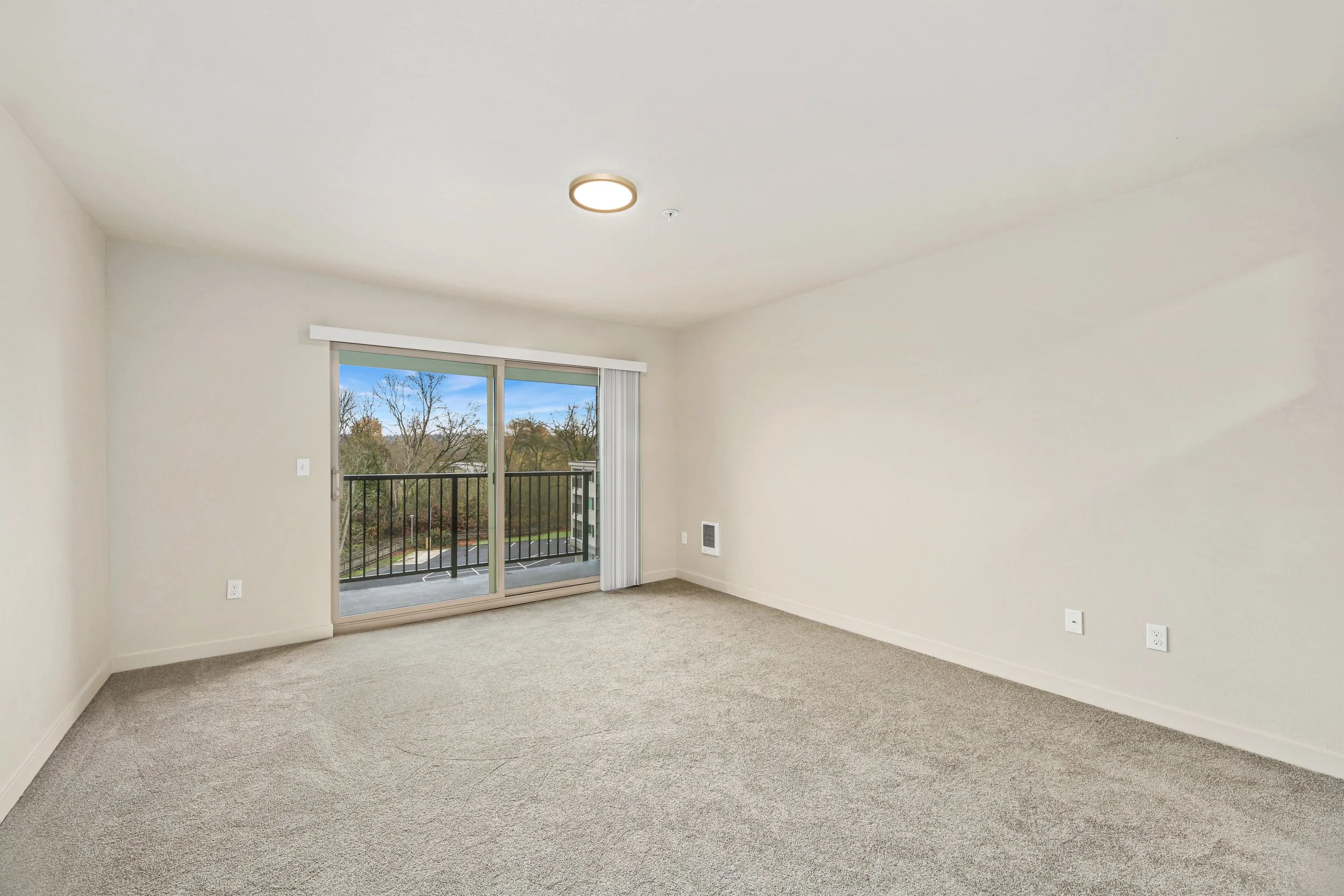 Empty living room with beige carpet, off-white walls, sliding glass door leading to a balcony, and trees outside.