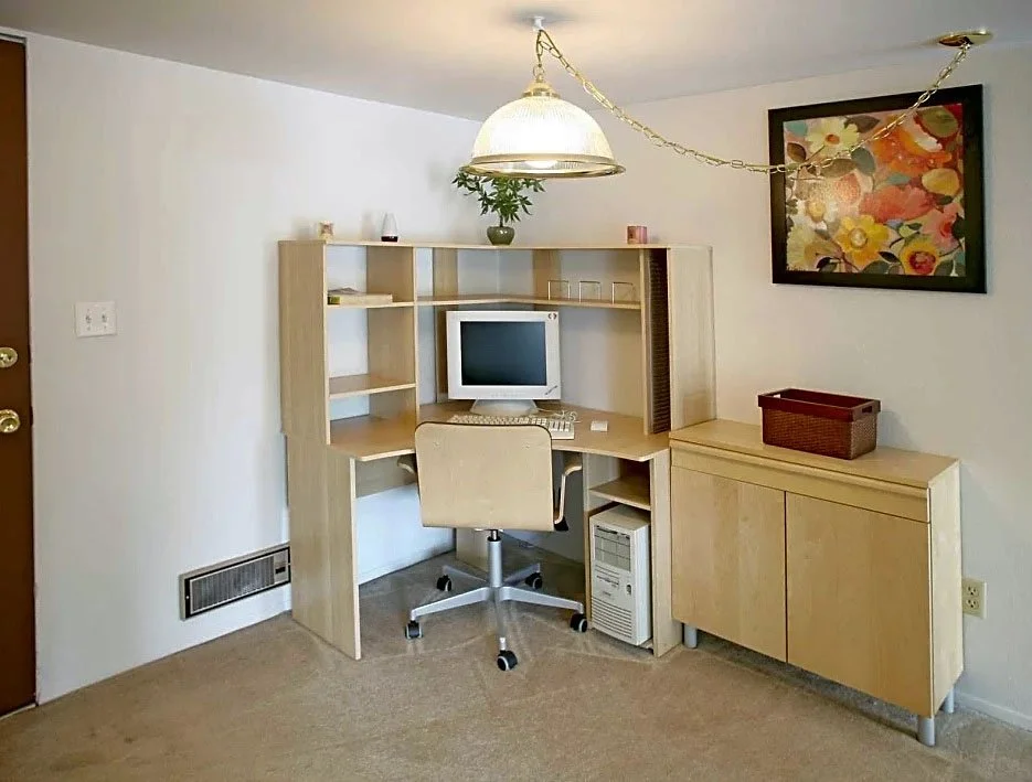 A small home office with a wooden desk and hutch, a beige rolling chair, a desktop computer with monitor, keyboard, and tower, a wooden cabinet with a basket on top, and a colorful floral painting on the wall. A hanging light fixture is also visible.