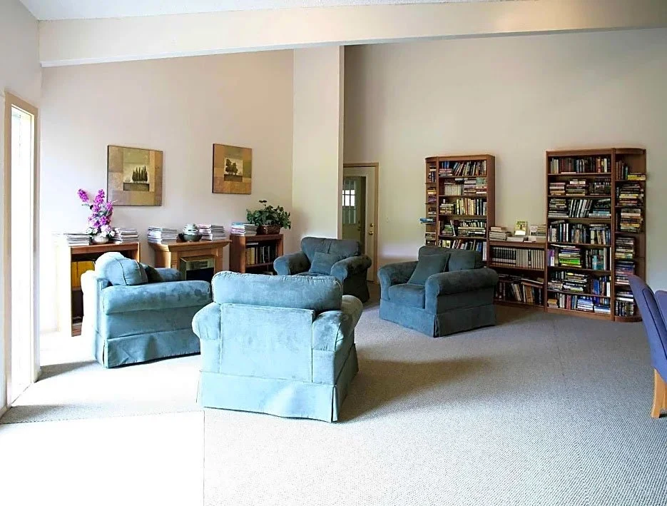 Living room with blue armchairs, wooden bookshelves filled with books, a side table with flowers and a plant, and artwork on the walls.
