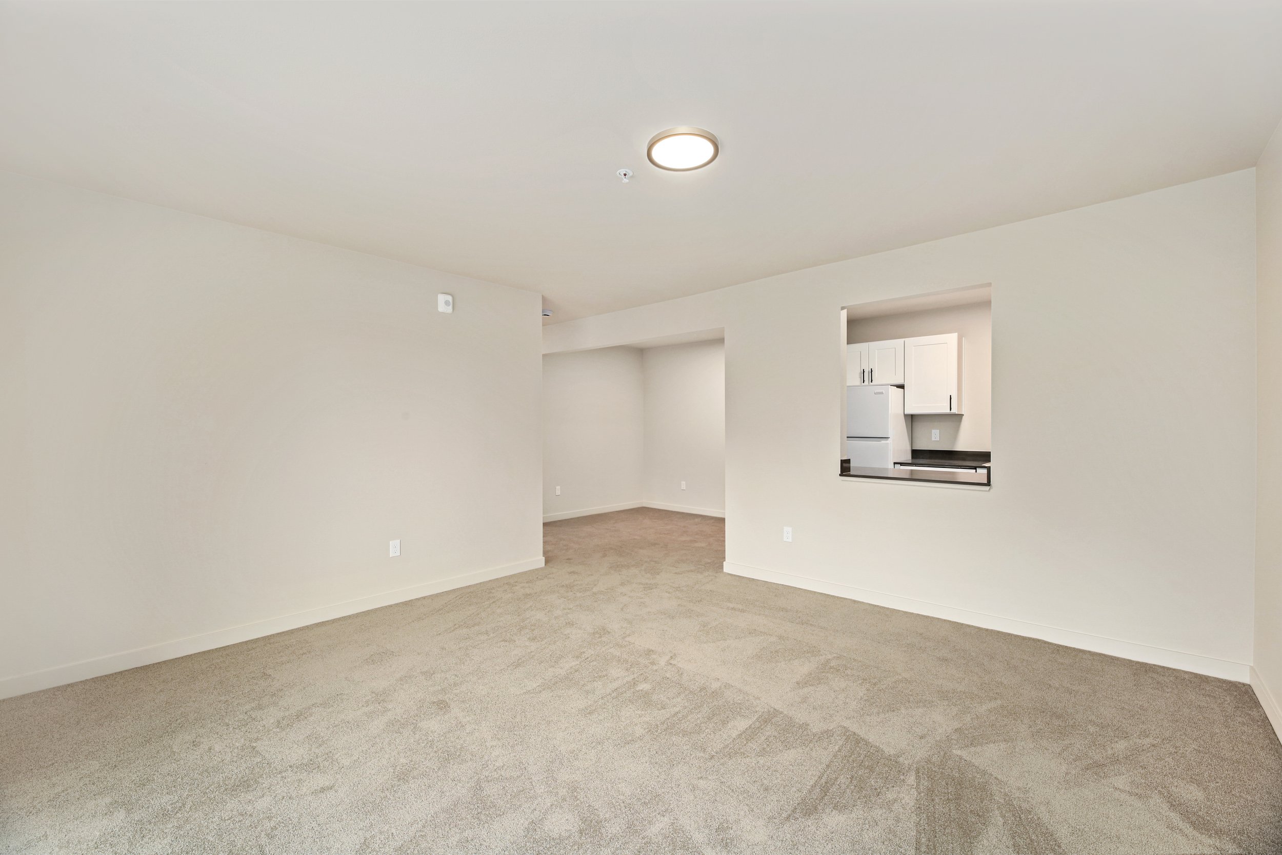 Empty living room with beige carpet, white walls, and open view to kitchen with white cabinets and refrigerator.