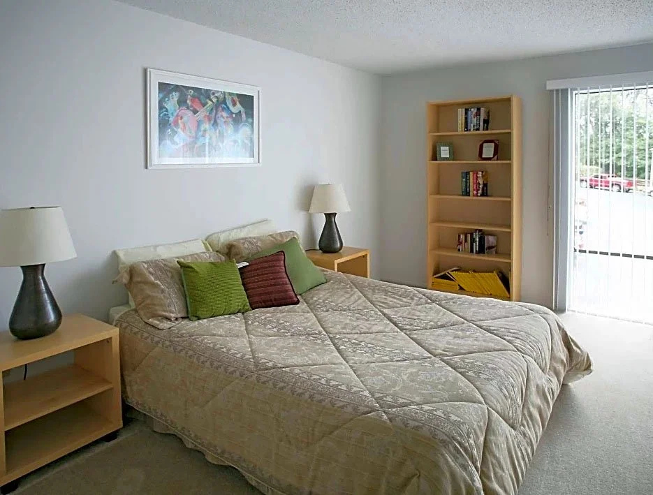 A bedroom with a queen-sized bed, beige bedspread, and three decorative pillows, flanked by two wooden nightstands with lamps. There is a wooden bookshelf near a sliding glass door, and a colorful framed picture hangs on the white wall above the bed.